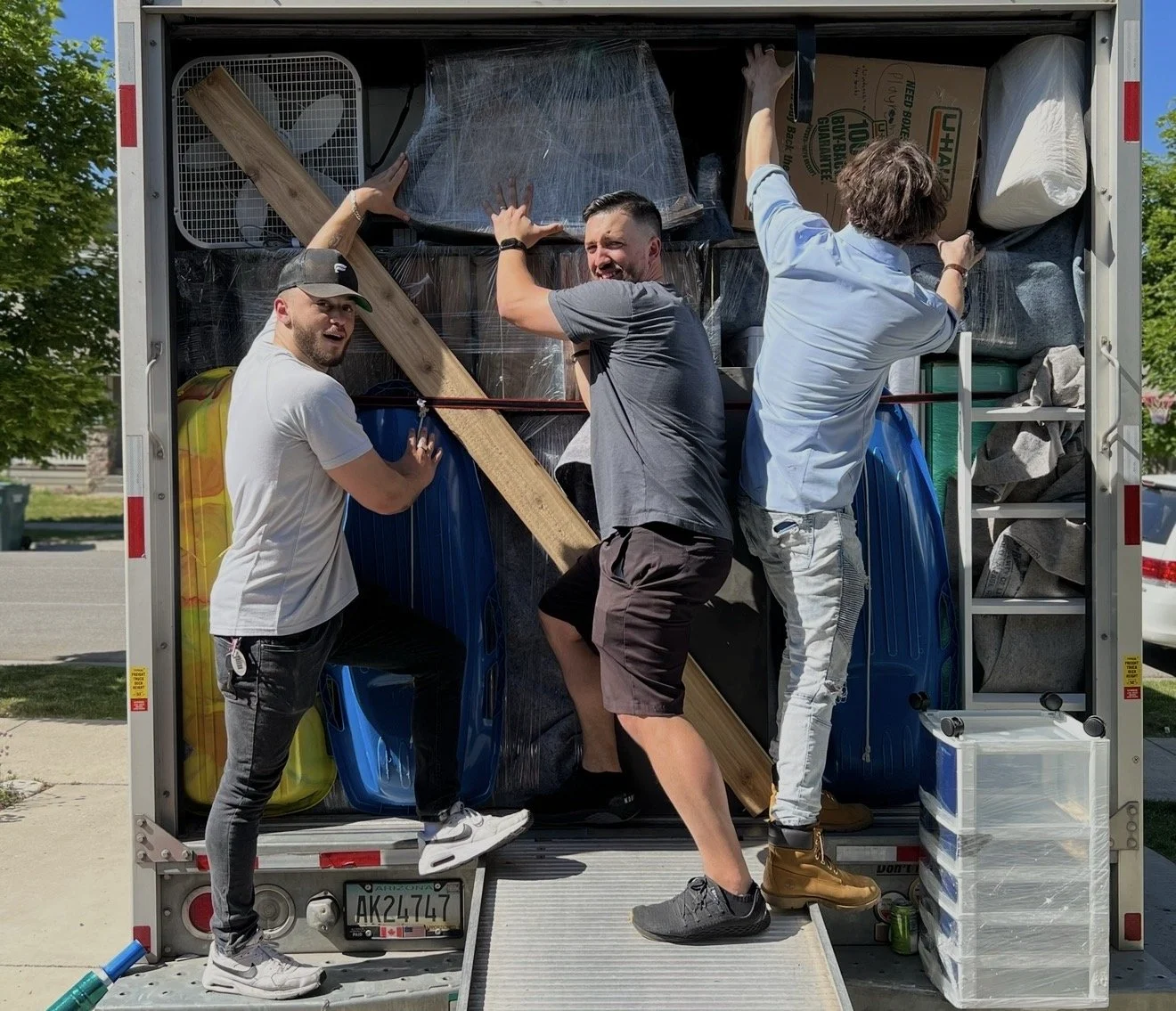 Three men are inside a moving truck loaded with various household items, including furniture, boxes, and a fan. They are working together to unload or organize the items.