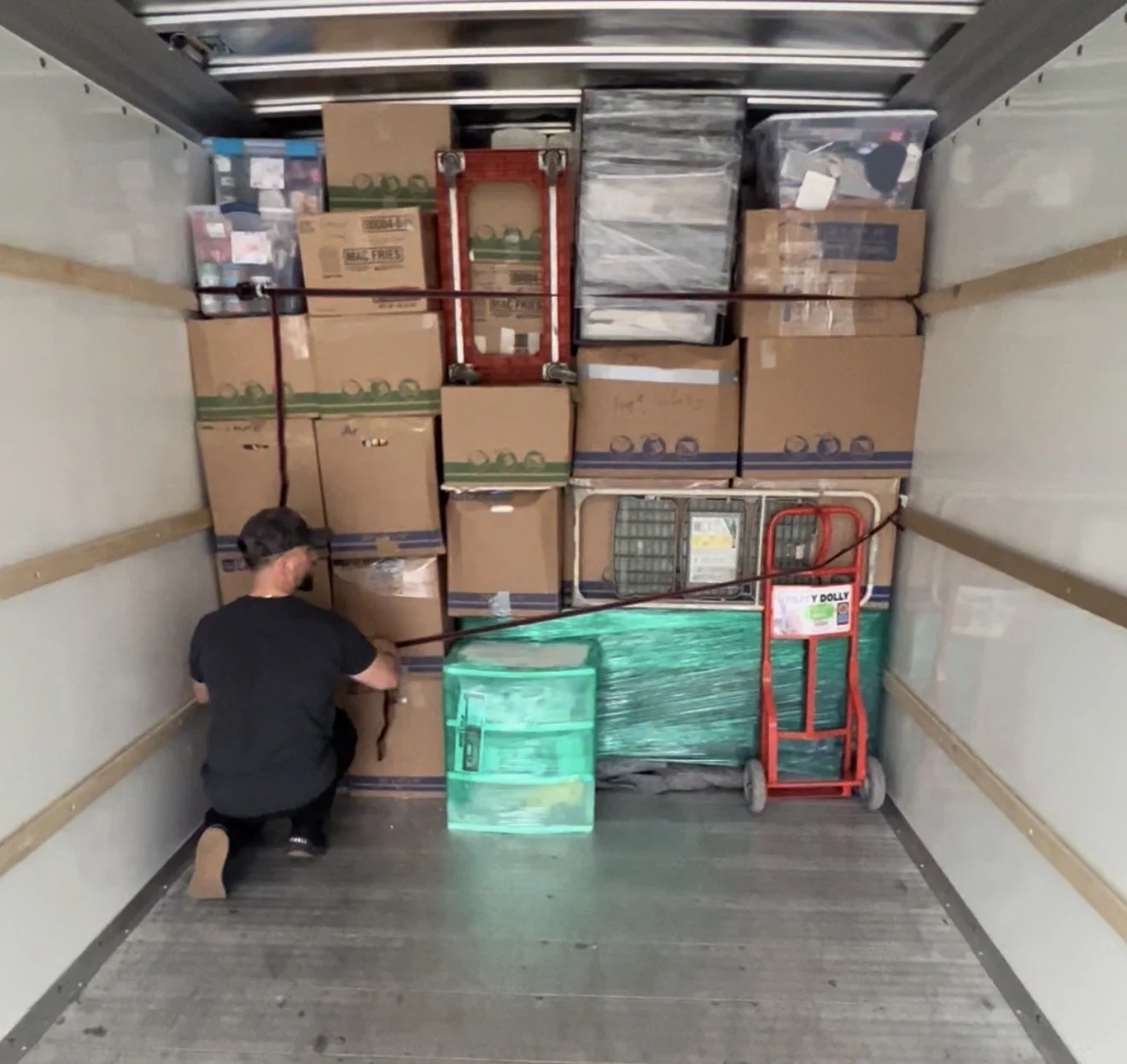 A person kneeling inside a moving truck packing boxes and plastic-wrapped pallets, with a red hand truck and various supplies.