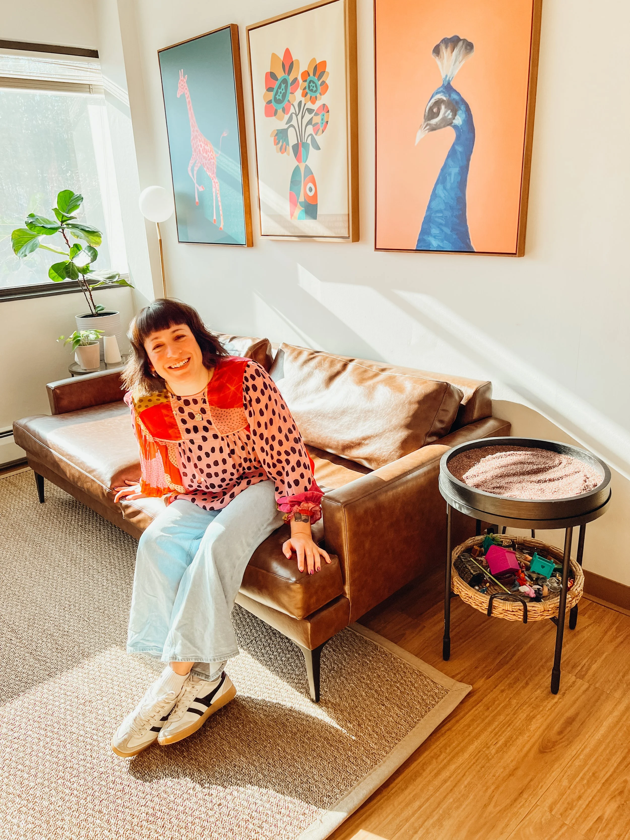 A woman sitting on a brown leather sofa in a living room with colorful artwork on the wall, a potted plant on a side table, and a tray with toys on a small table.