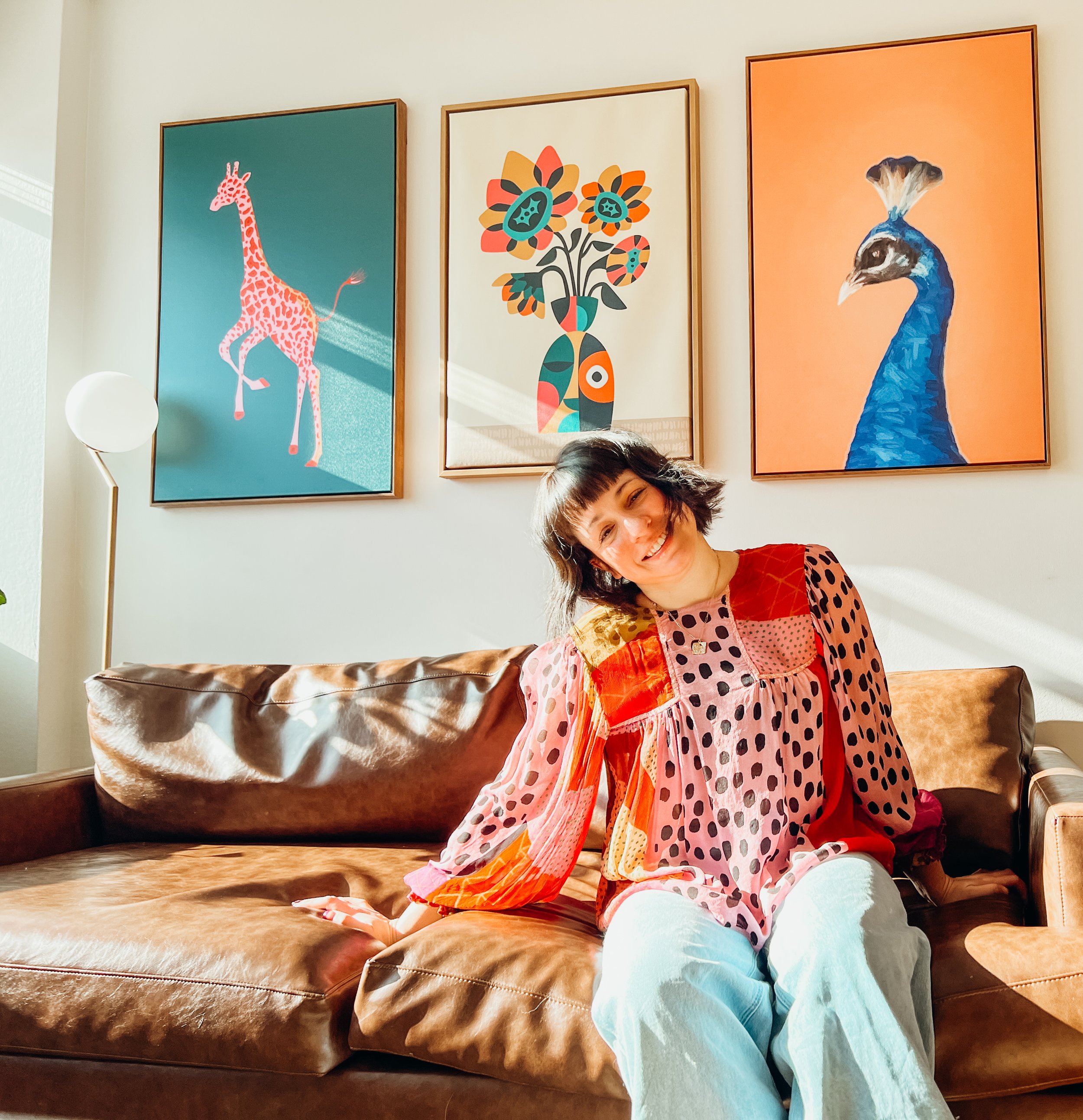A woman with short dark hair and a multicolored polka dot blouse sitting on a brown leather sofa in a well-lit room, with three framed colorful animal and plant artwork on the wall behind her.
