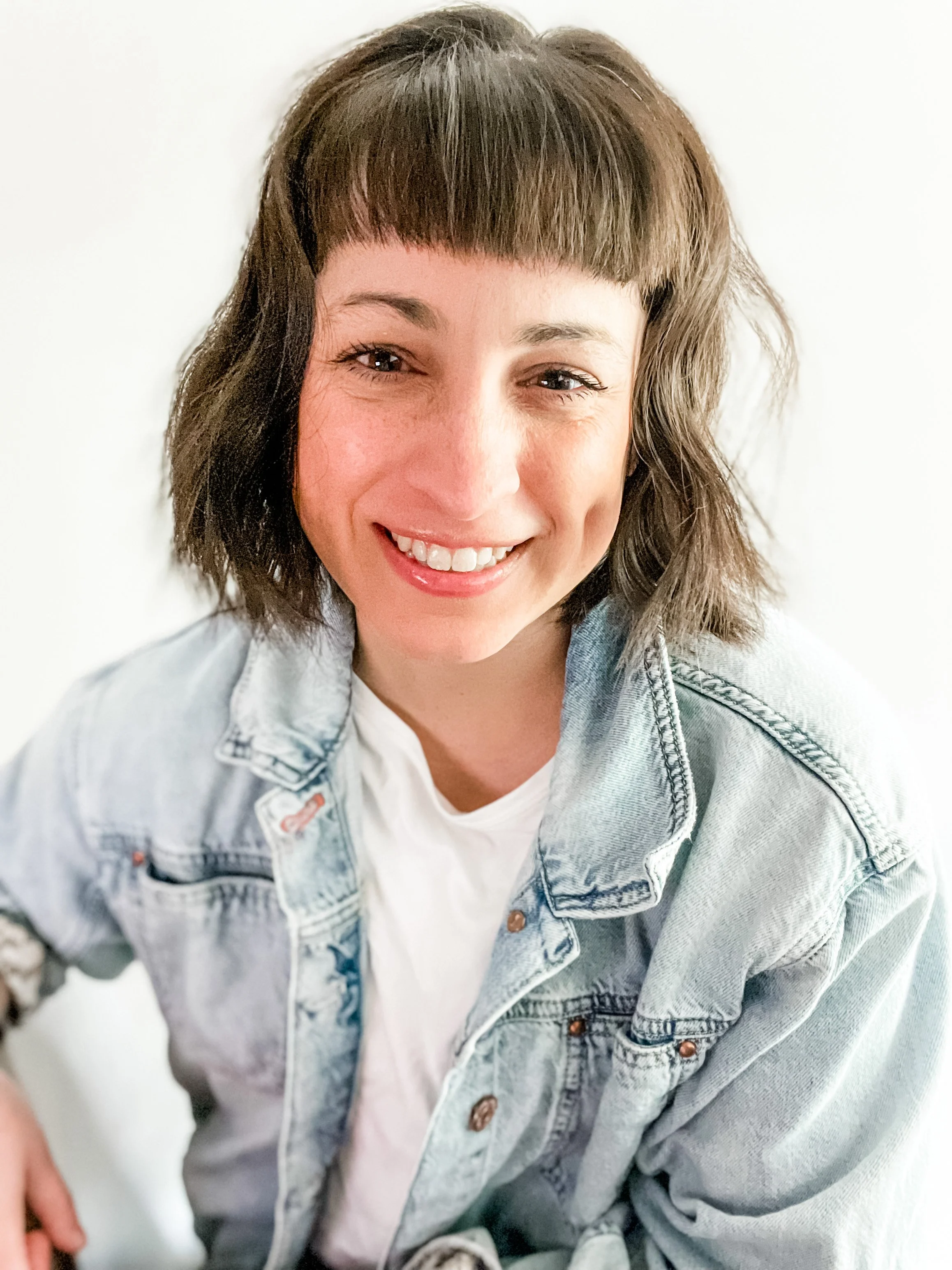 A young woman with short brown hair and bangs, smiling while looking into the camera, wearing a light blue denim jacket over a white shirt, standing against a plain white background.