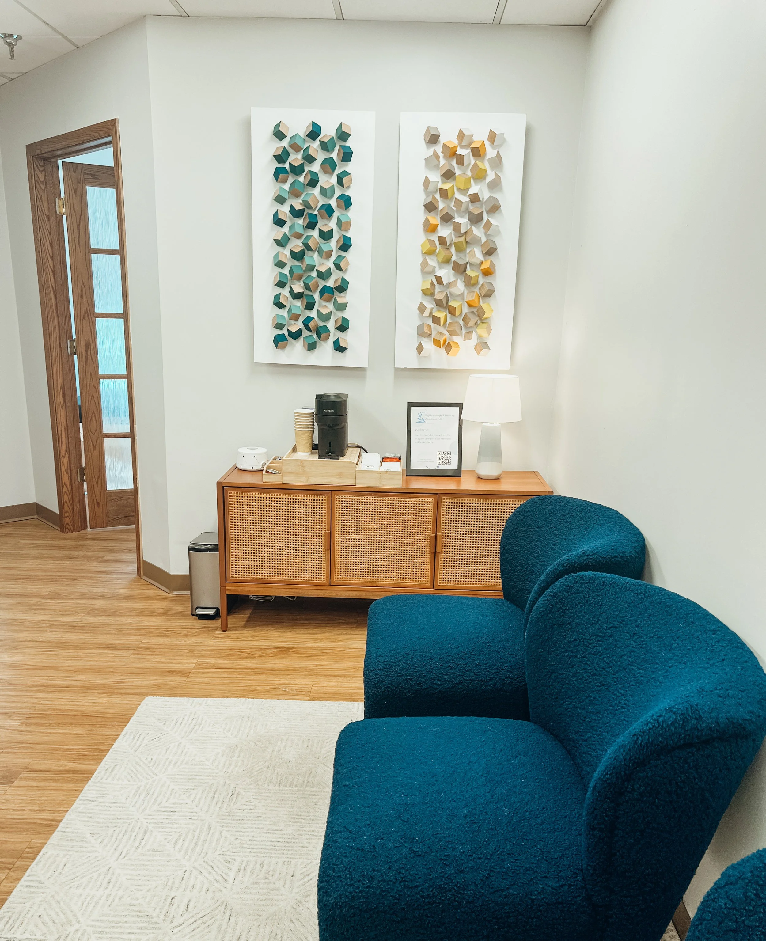 Waiting area with three blue textured chairs, a wooden cabinet with coffee supplies, and two abstract wall art pieces with geometrical shapes on a white wall.