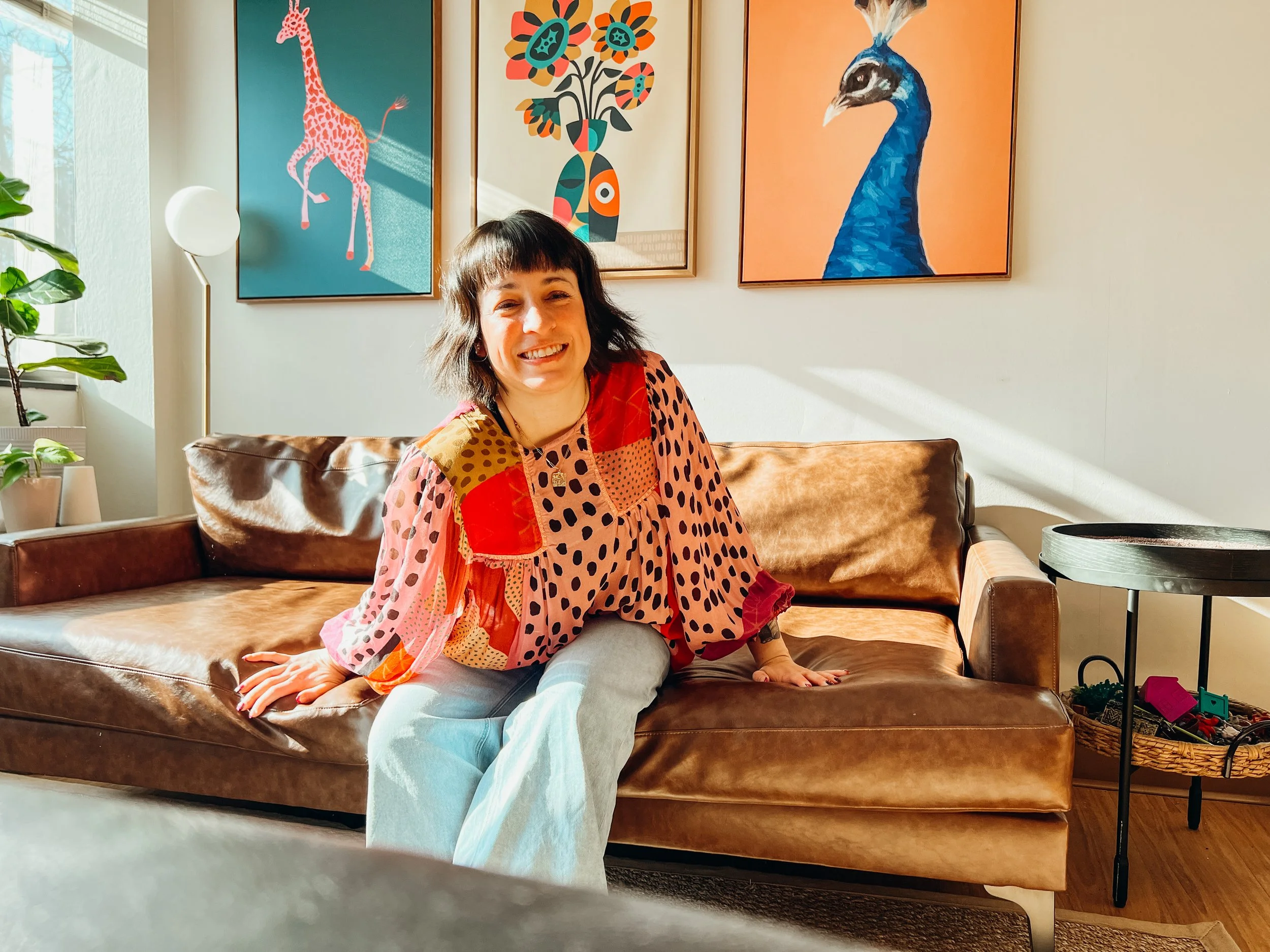 A woman with short dark hair sitting on a brown leather sofa, smiling, wearing a colorful blouse with polka dots and patches, in a bright living room with three colorful animal portraits on the wall behind her.