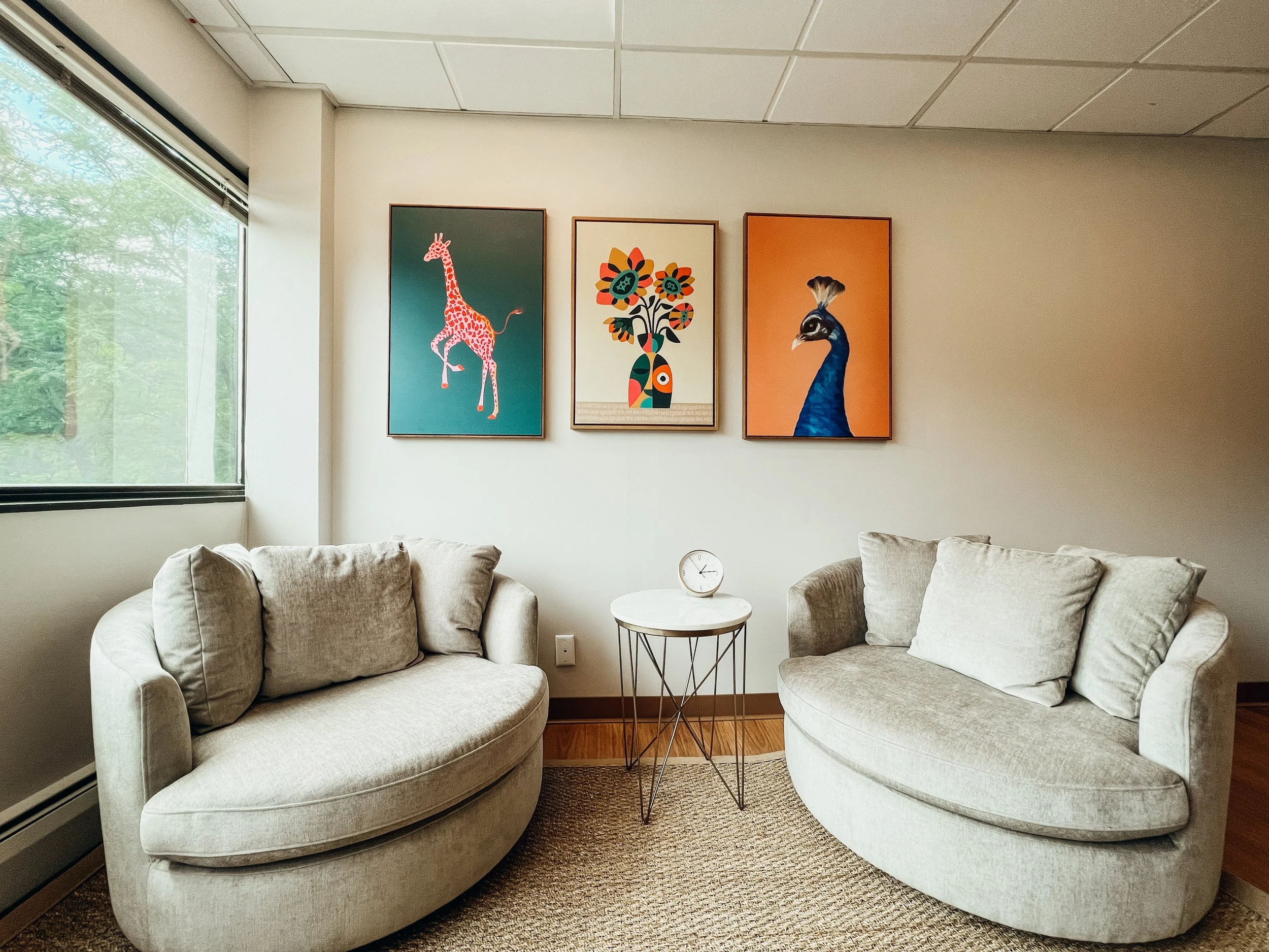 A cozy waiting area with two beige armchairs and a small round white table with a clock on it. The wall behind has three colorful animal and floral prints, with a large window on the left showing green trees outside.