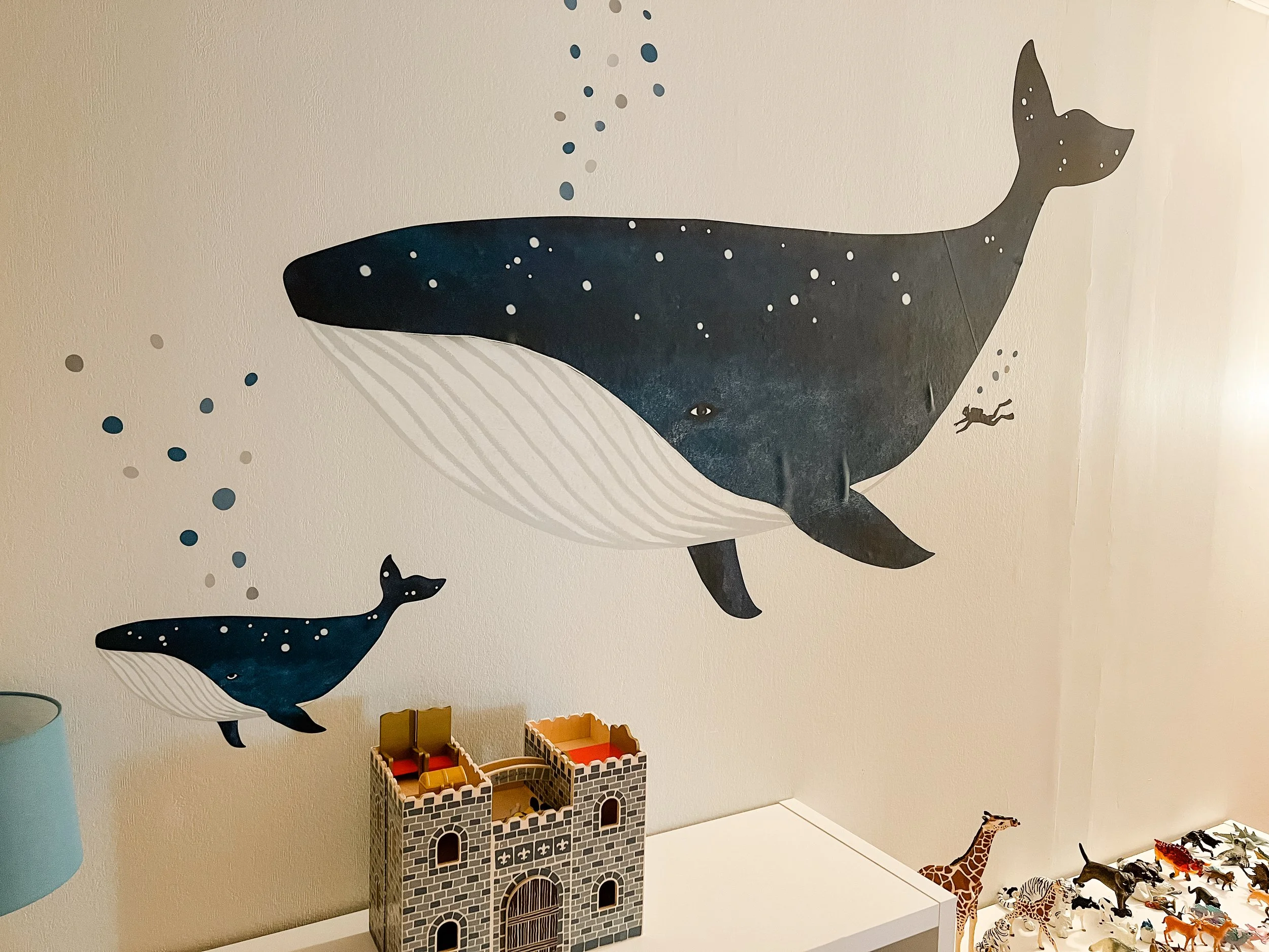 Wall mural of a whale with starry night design, with a smaller whale nearby and a diver silhouette, seen above a toy castle and giraffe figurine, and toy animals on a white surface.