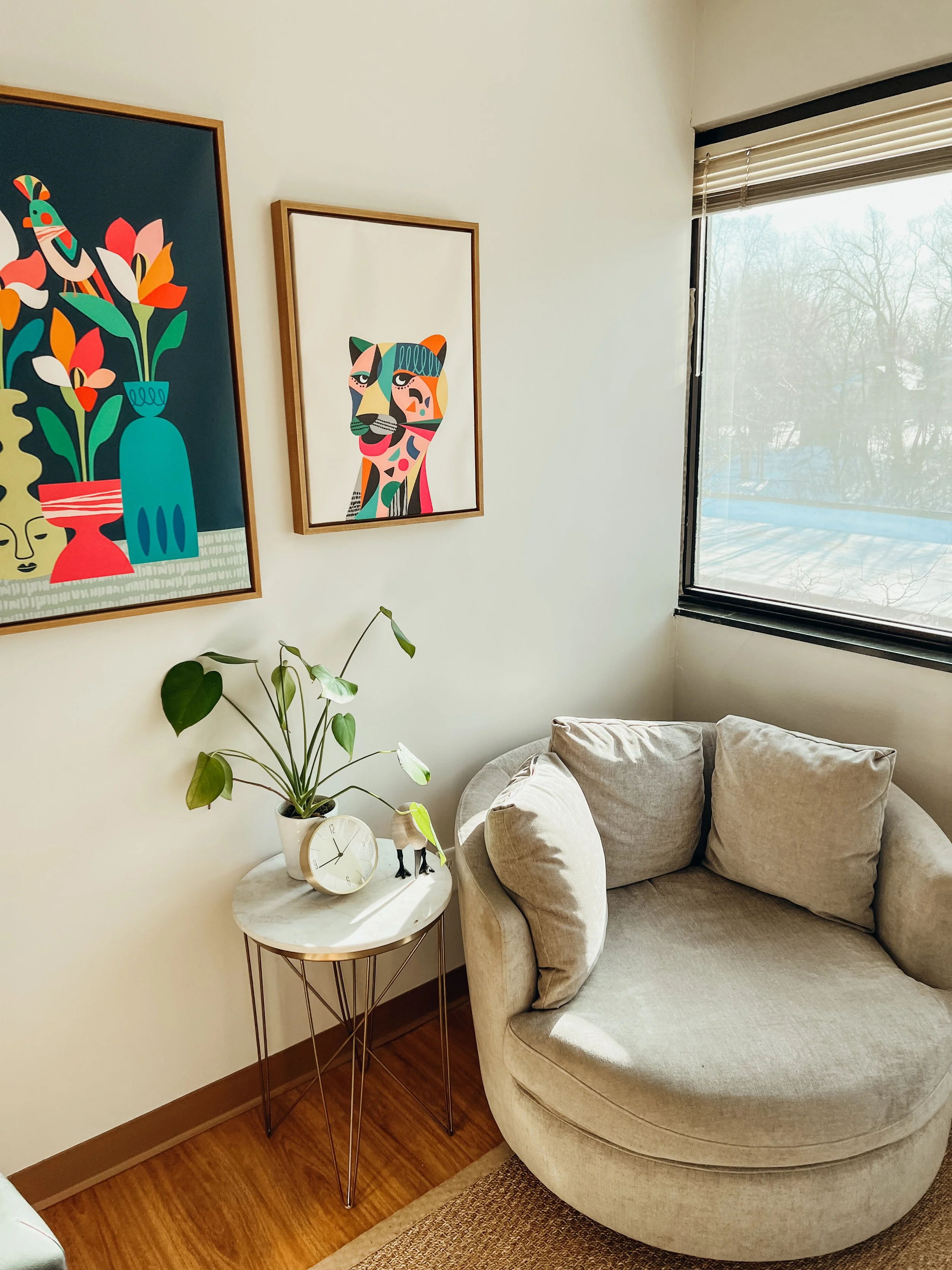 Cozy living room corner with a round beige sofa, a small marble-top side table with a clock, a plant, and small decorative items, and framed colorful modern art on the wall near a large window.