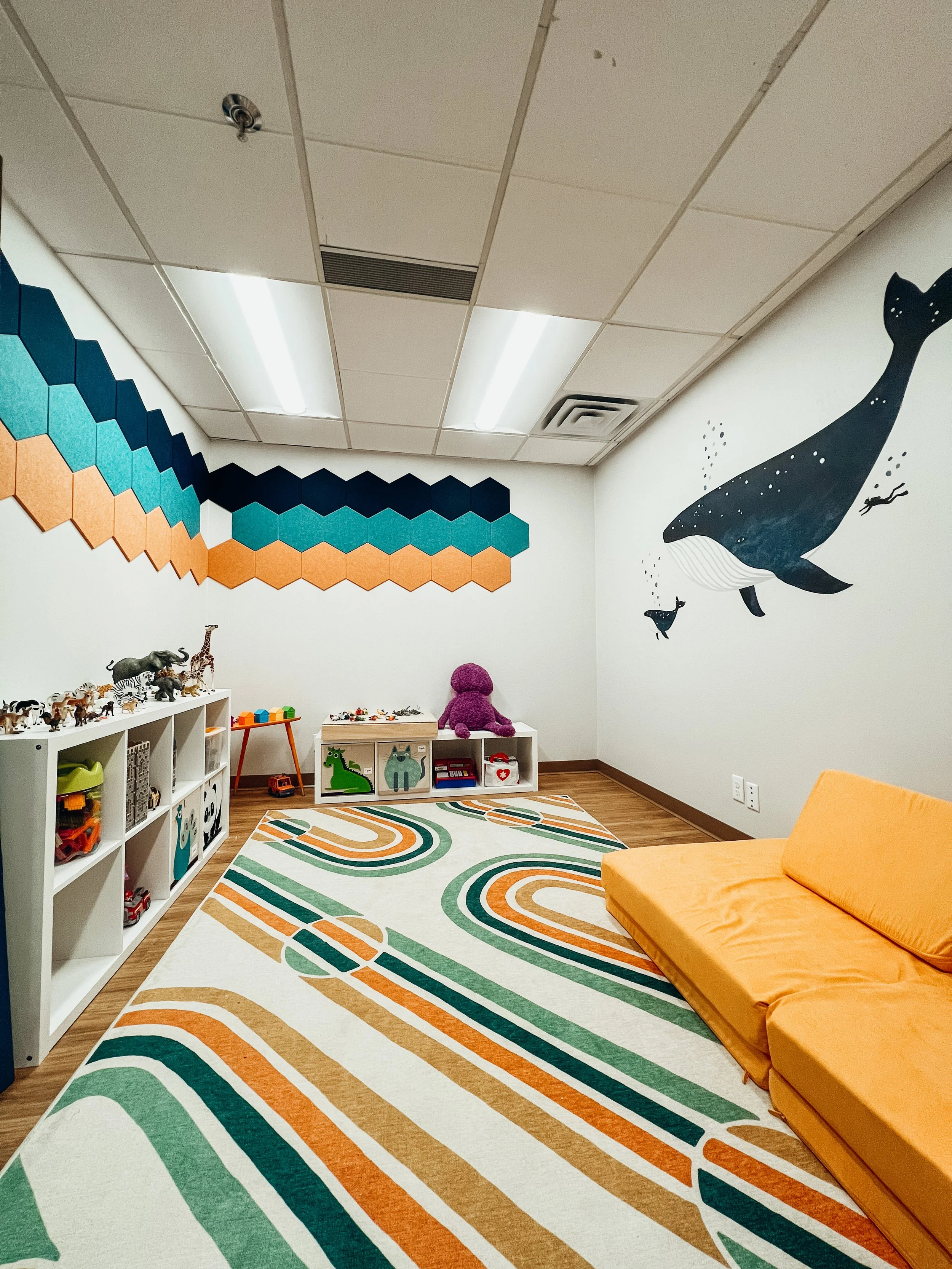 Colorful children's playroom with animal toys, a patterned rug, a bright orange couch, and wall murals of a whale and geometric shapes.