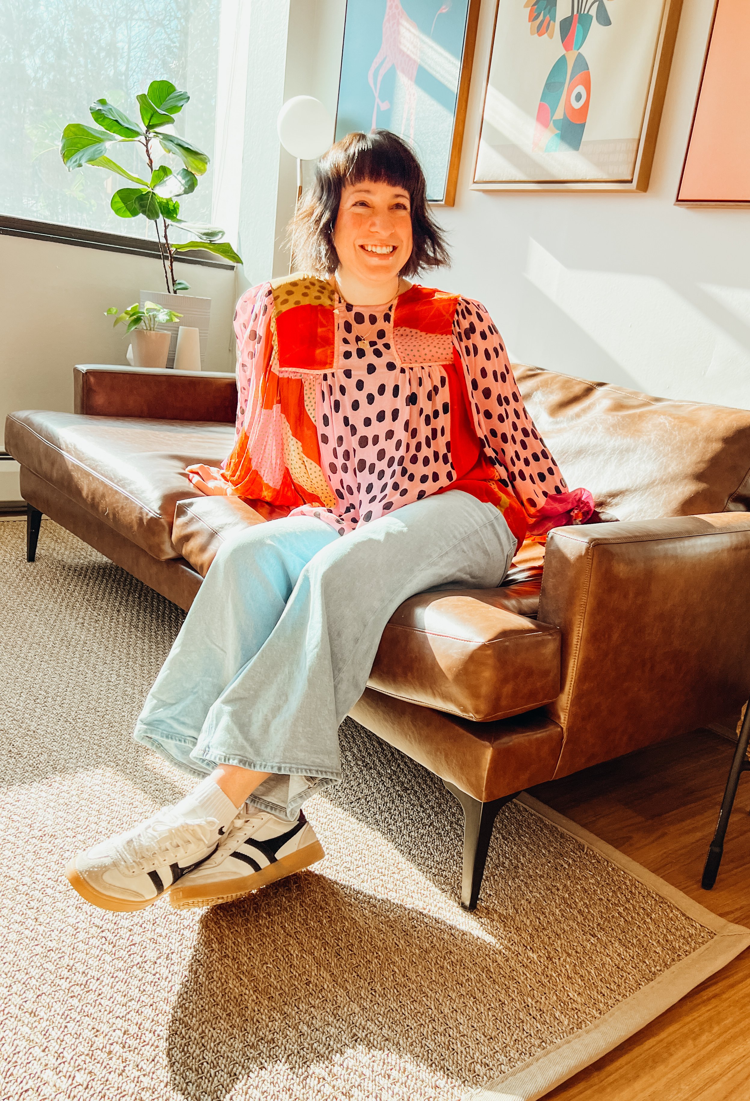 A woman with dark, shoulder-length hair and bangs, smiling and sitting on a leather sofa in a brightly lit room with plants and colorful framed artwork on the wall.