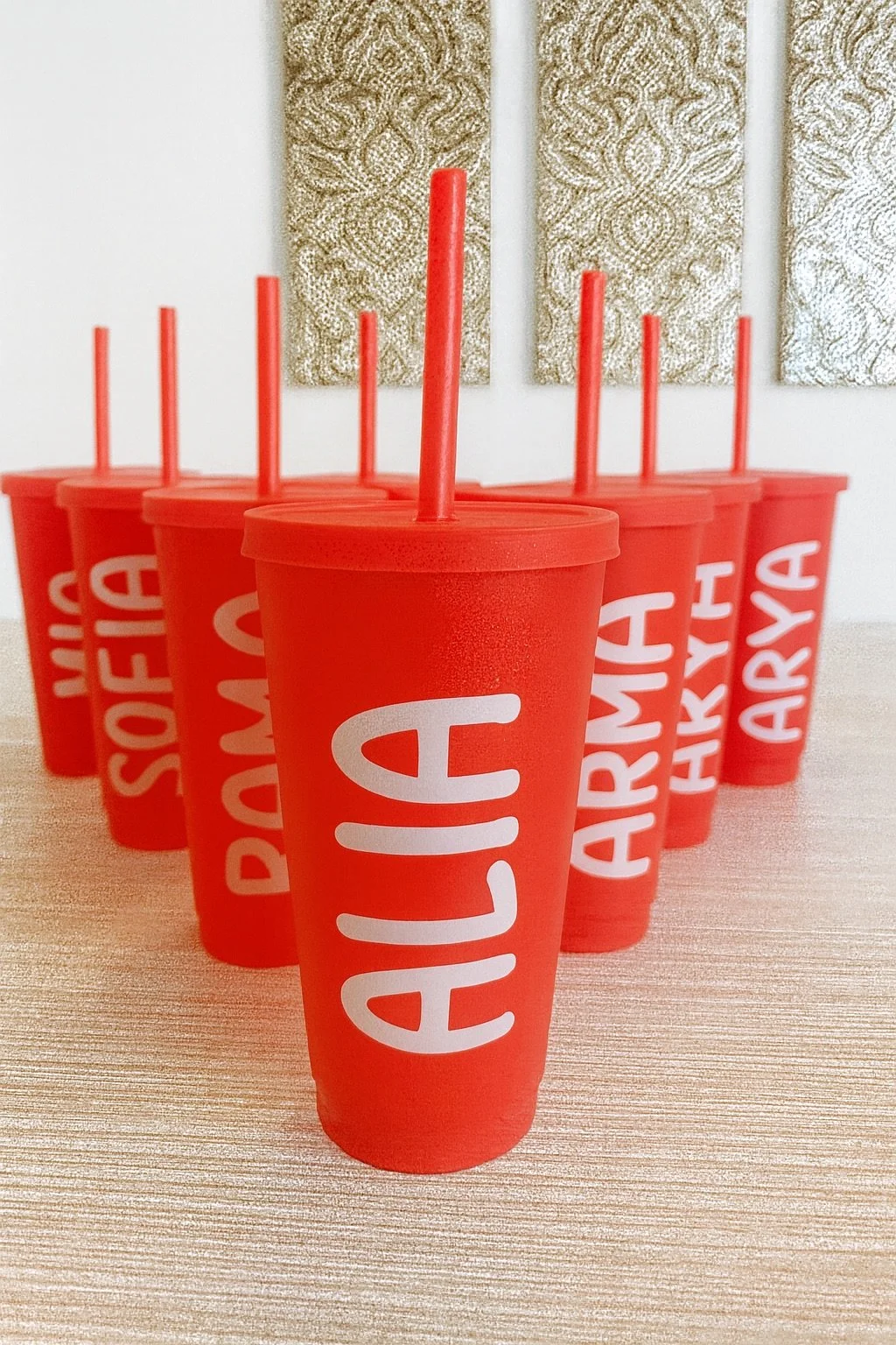 A row of red cups with white straws and the word "ALAI" printed on them, arranged on a wooden surface in front of a textured wall.