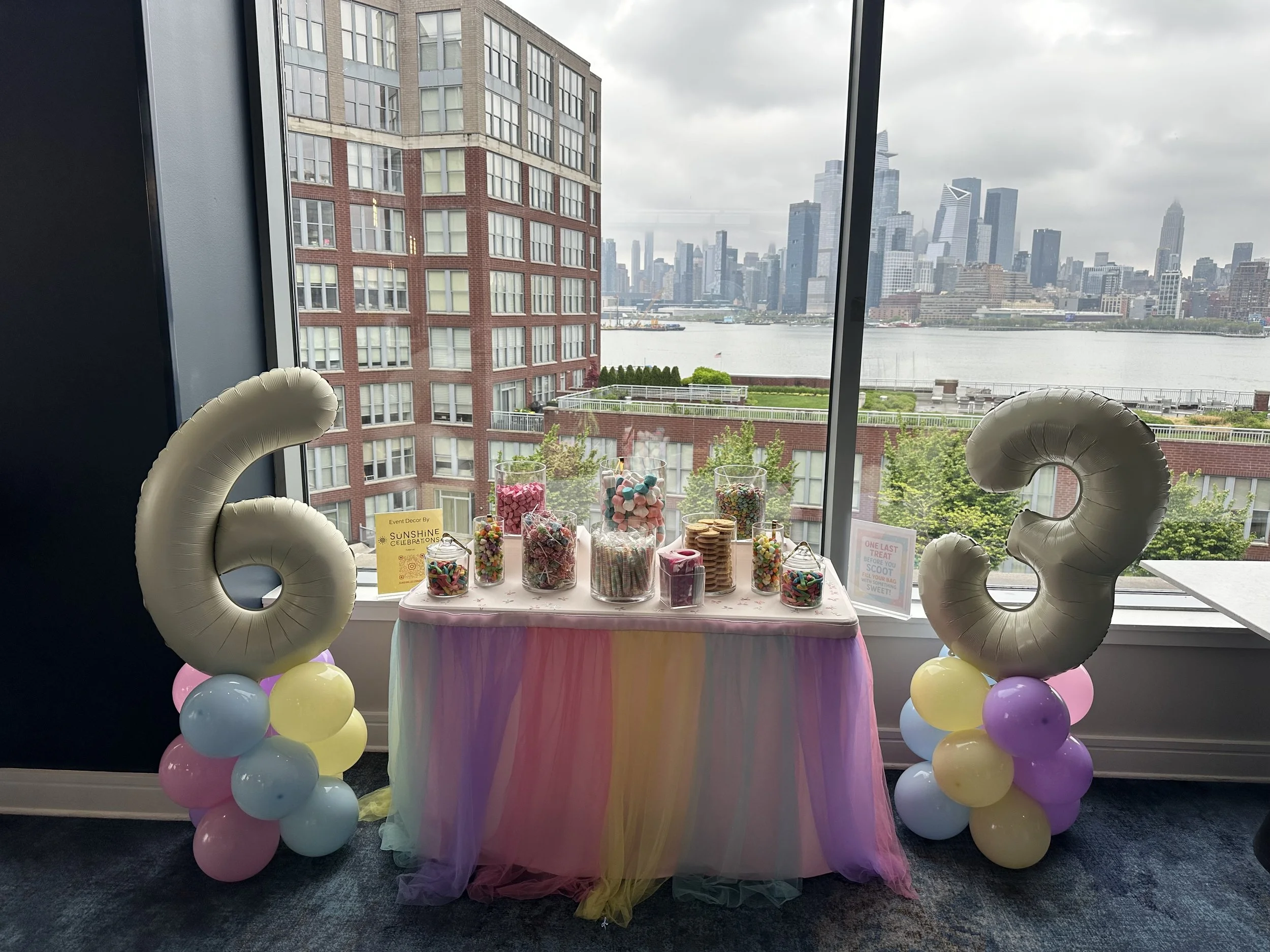 Candy table decorated with pastel balloons, a rainbow tulle skirt, and large balloon numbers '6' and '3' in gold, with city skyline and river view through large windows in background.