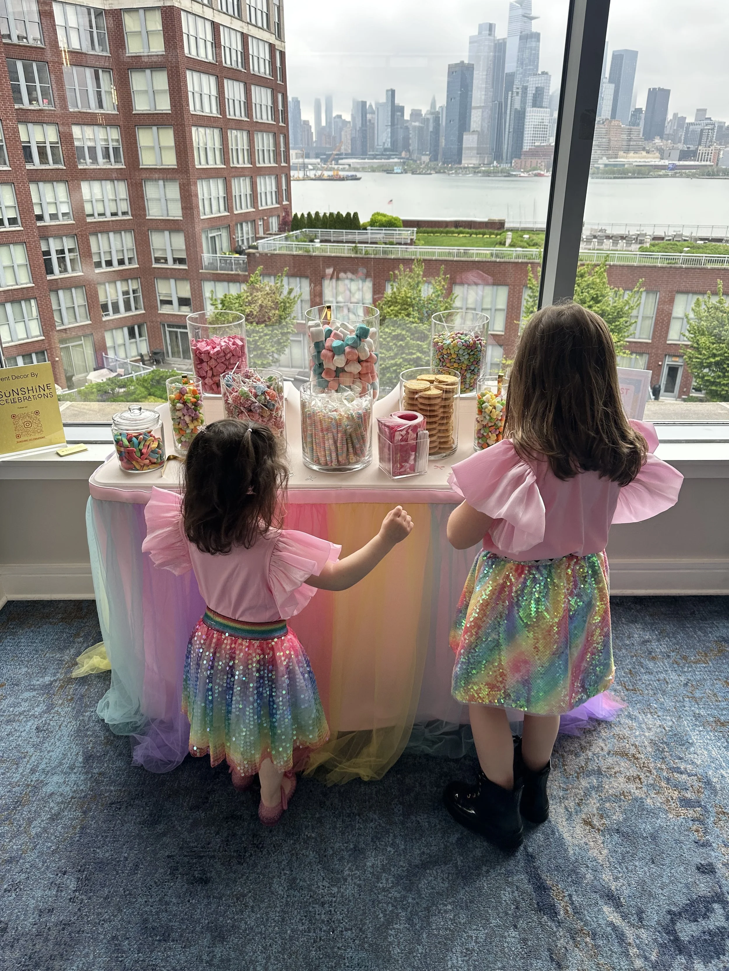 Two young girls in colorful rainbow skirts and pink shirts standing in front of a candy table decorated with a pastel pink and yellow fabric, overlooking a city skyline through large windows.
