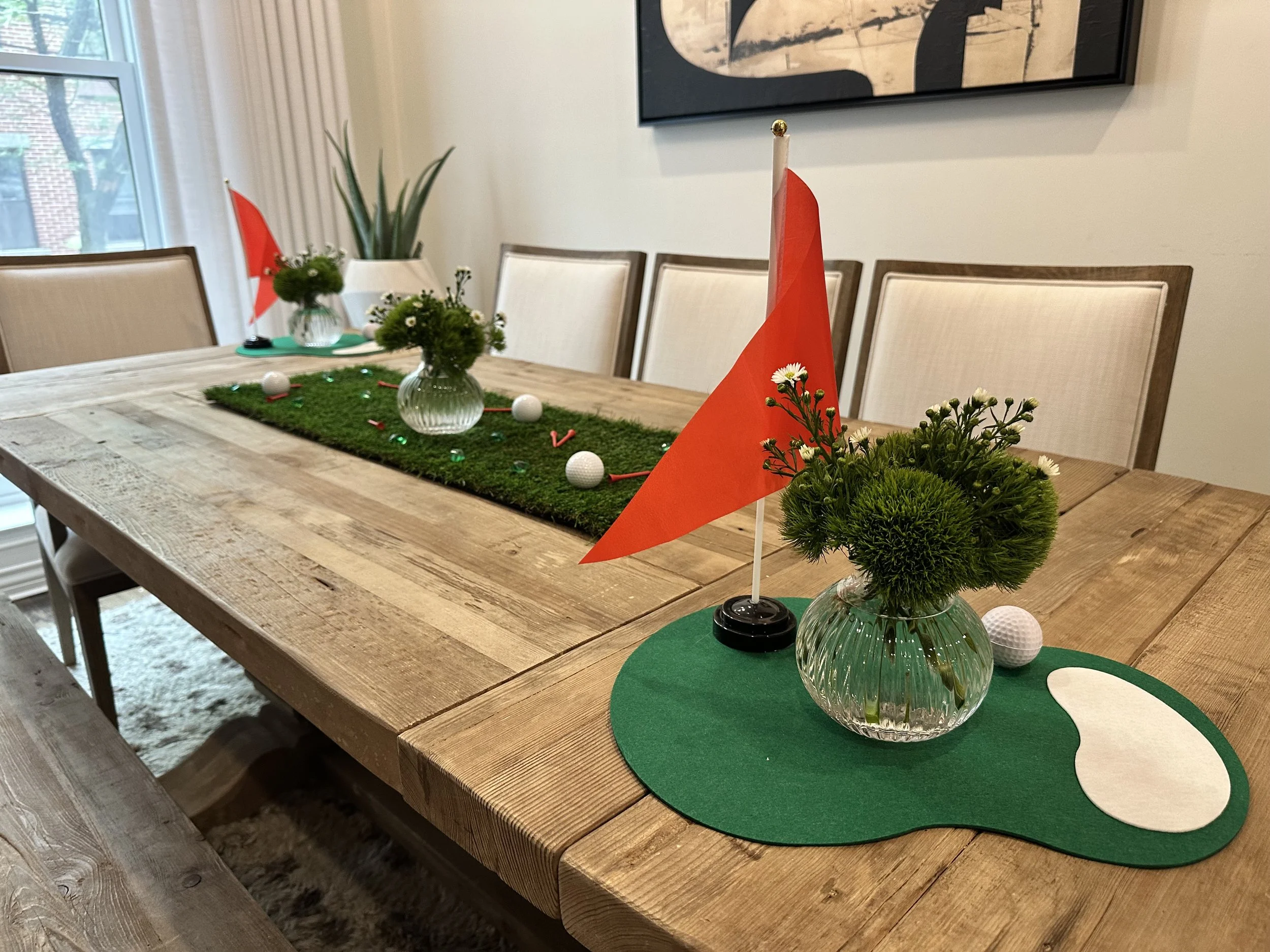 A dining table decorated for Valentine's Day with a green felt placemat, a glass vase with white flowers, and a small orange flag. Additional similar decorations with vases, flags, and plants are on the table.