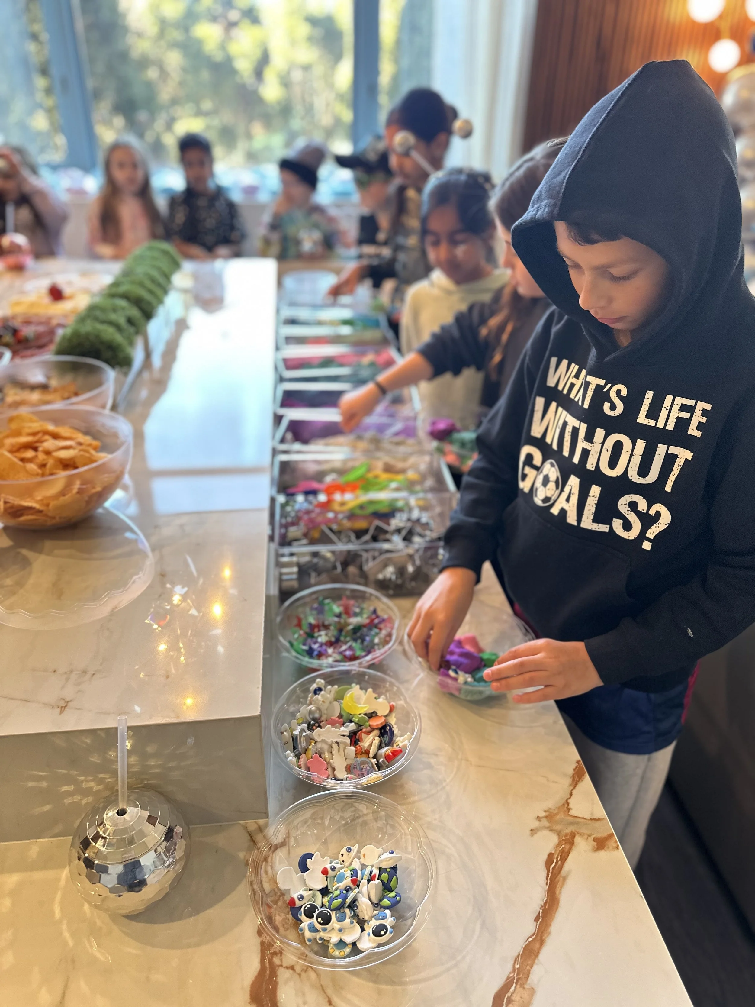 Children at a table enjoy choosing colorful decorations, such as stickers, for a craft or activity in a brightly lit indoor space with large windows and trees outside.