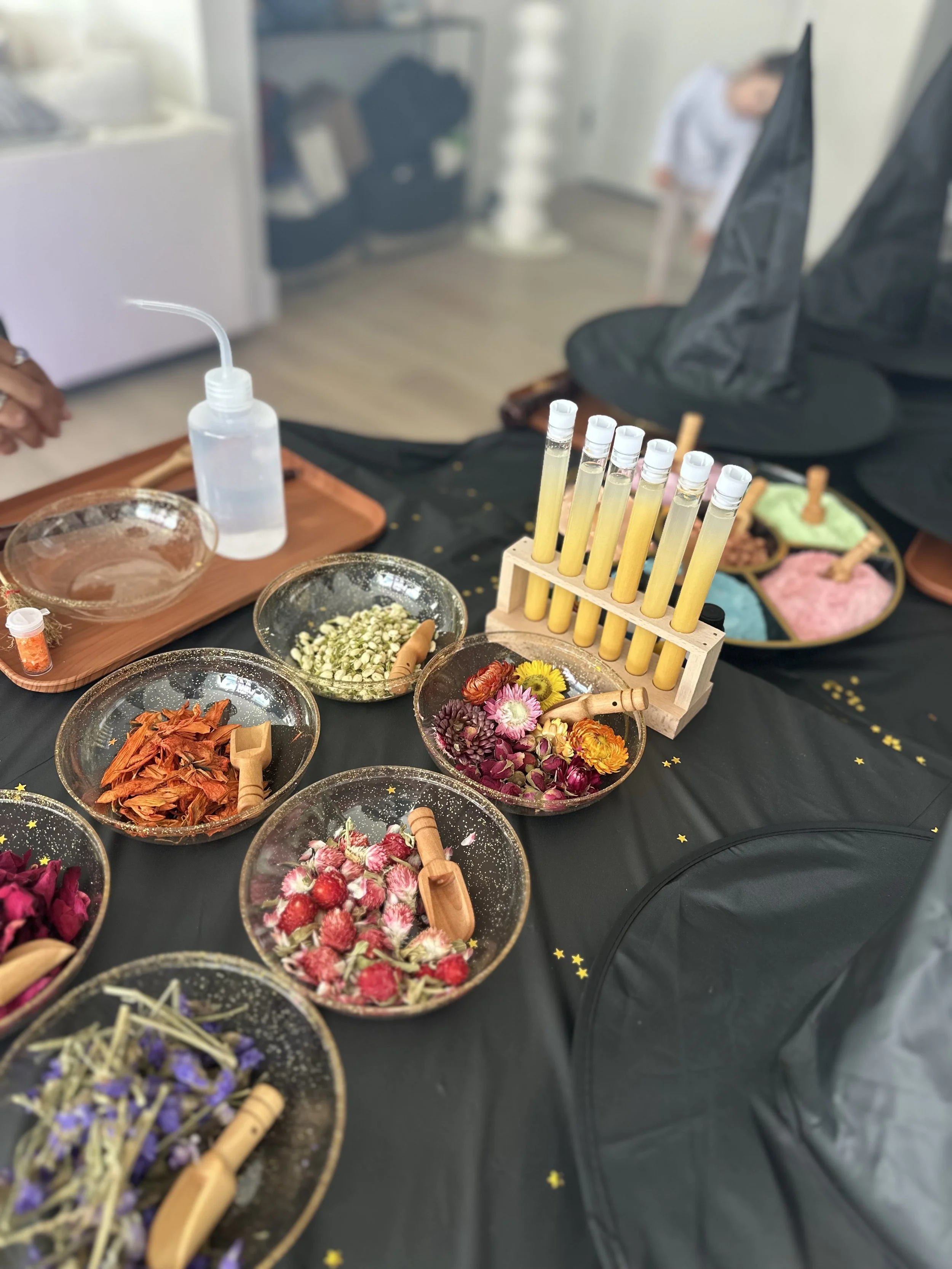 Display of colorful dried flowers, test tubes with yellow liquid, and containers with dyed powders on a black table, possibly for a flower dyeing or herbal infusion activity.