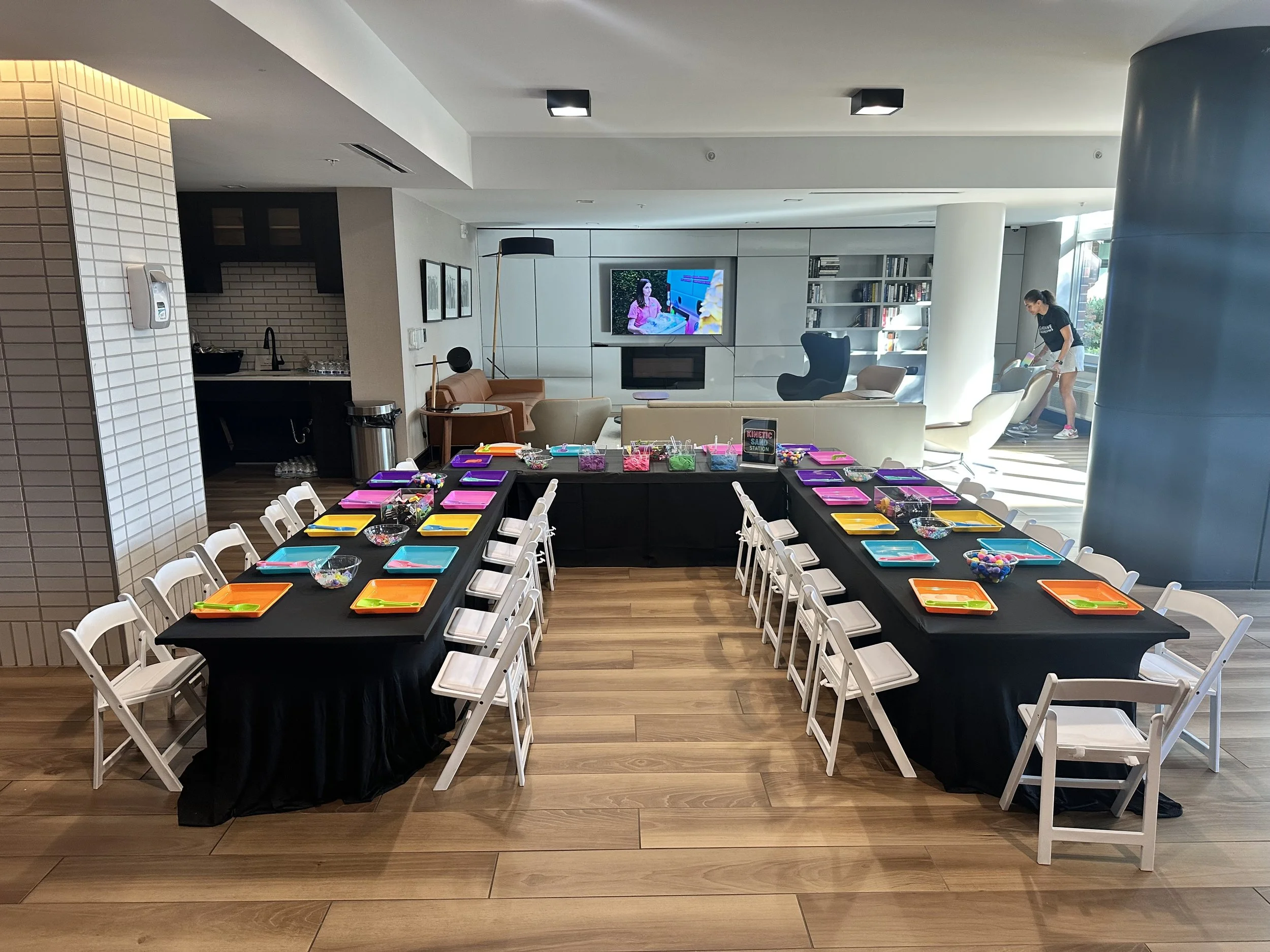 A room with tables decorated for a children's party, featuring colorful plates and bowls, and a background with modern furniture, a TV, and a person cleaning.