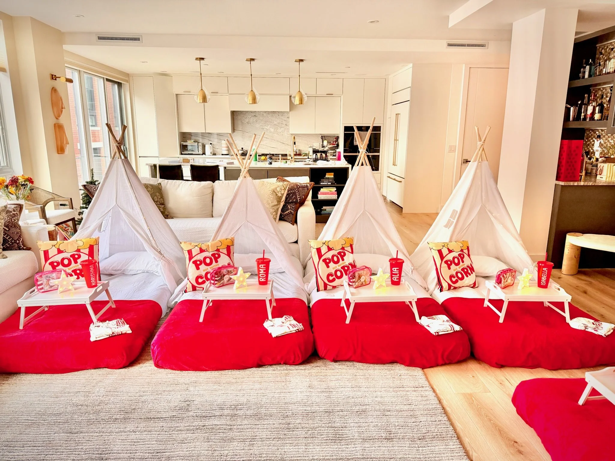 Four red cushions with white teepees on top, decorated for a birthday celebration, in a modern living room with a kitchen in the background.