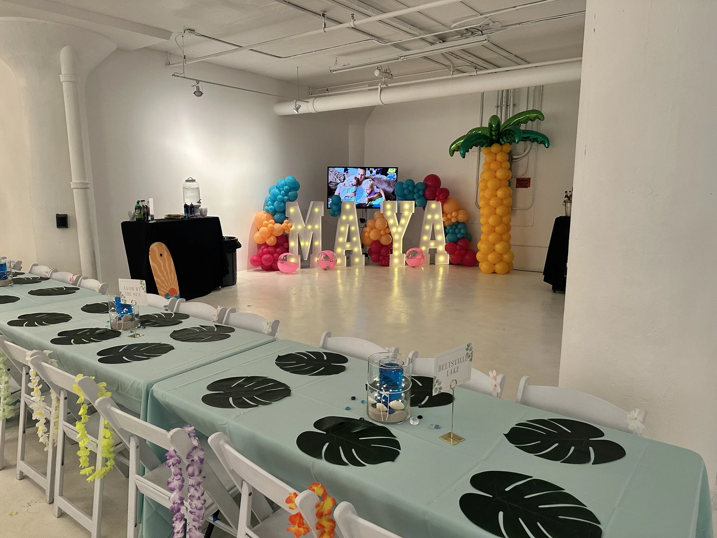 Party setup with a long table covered in a TikTok-themed tablecloth with black leaves, decorated with mini leis, and a backdrop with balloons shaped like a palm tree and large letter lights spelling 'MAYA' with a TV and colorful balloons in the backg