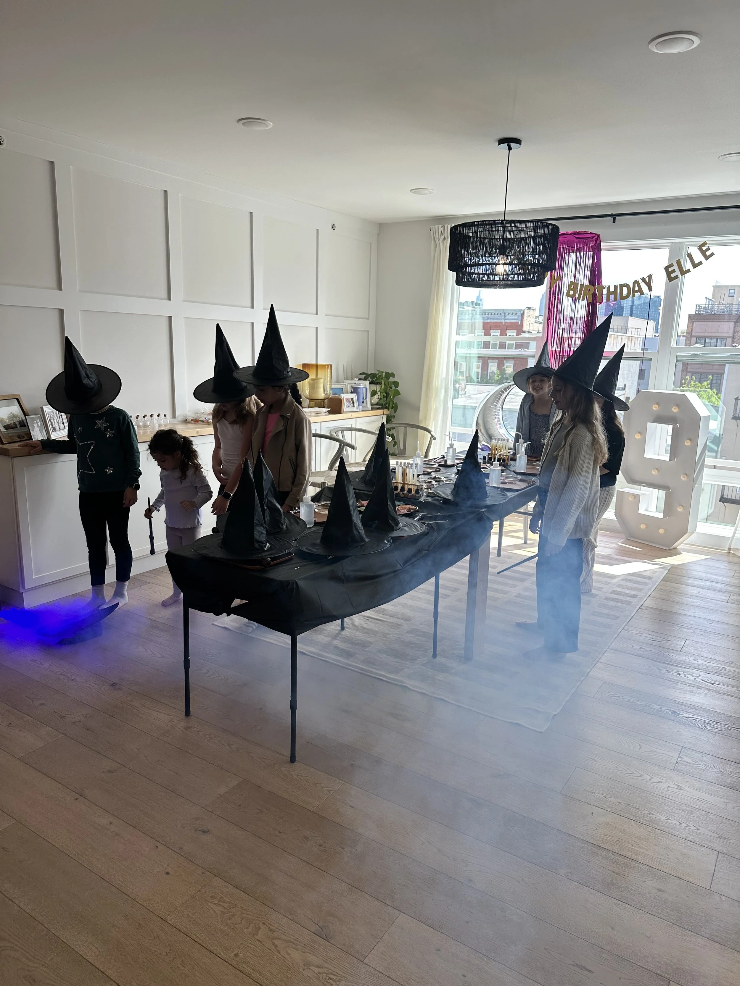Children wearing witch hats gathered around a decorated table with a Halloween theme, preparing for a celebration in a bright room with wooden floors and large windows.