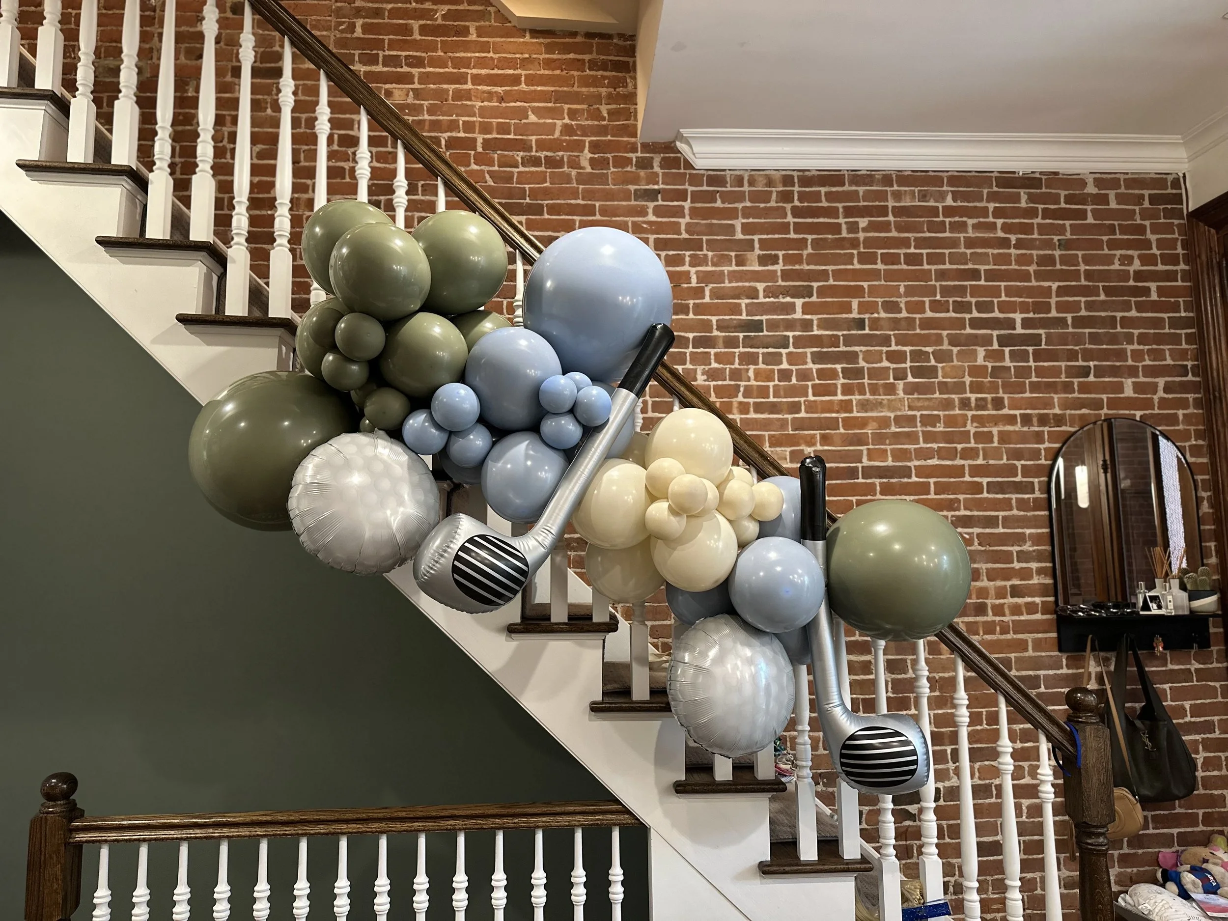 Decorative balloon arrangement on staircase railing with a mix of green, blue, white, and silver balloons, and two large inflatable microphones. The staircase has wooden steps and white spindles, with a brick wall background.