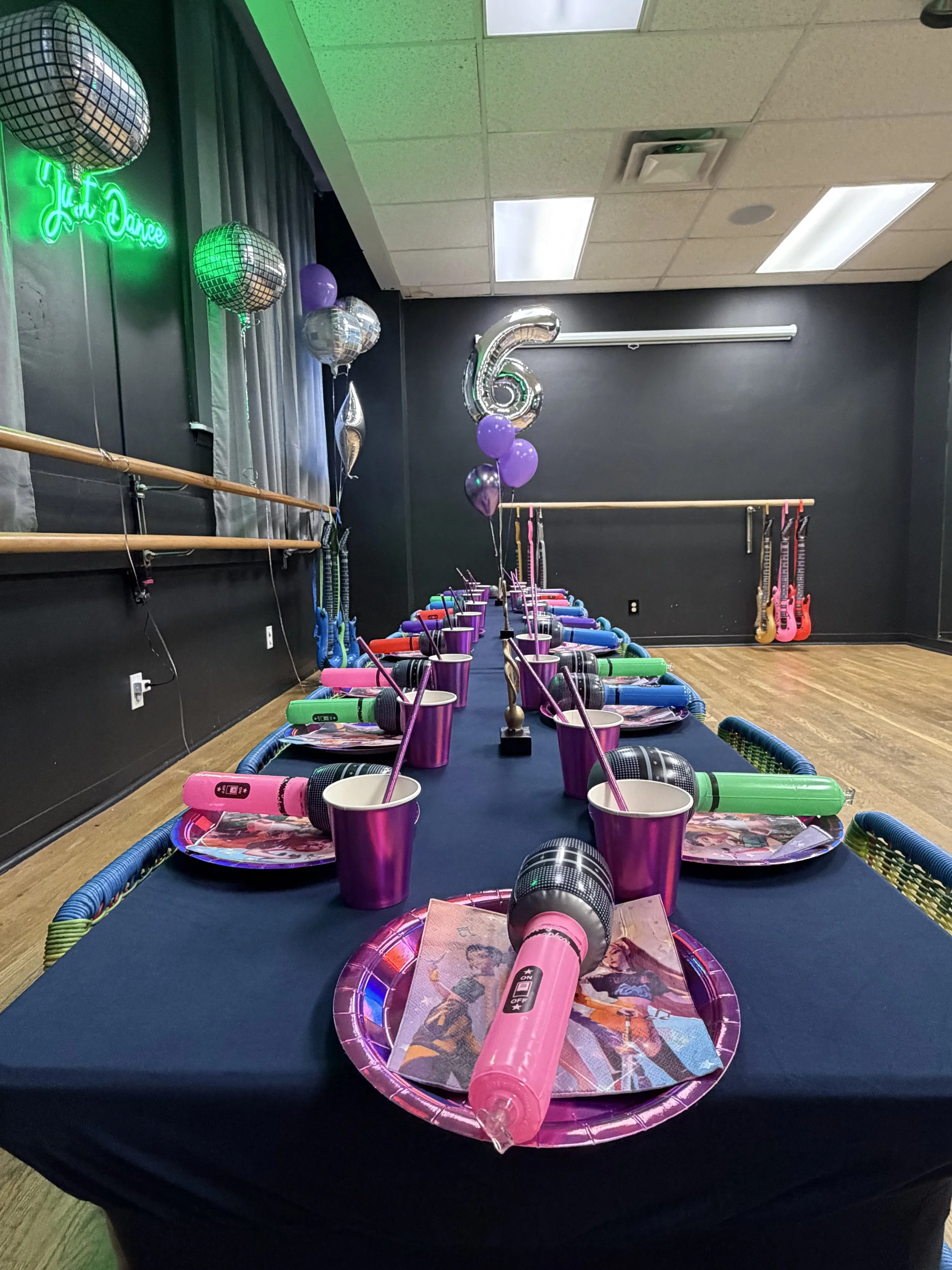 Decorated party room with a 6th birthday theme, featuring balloons, a long table with plates, cups, and microphones for karaoke, and pink guitars in the corner.