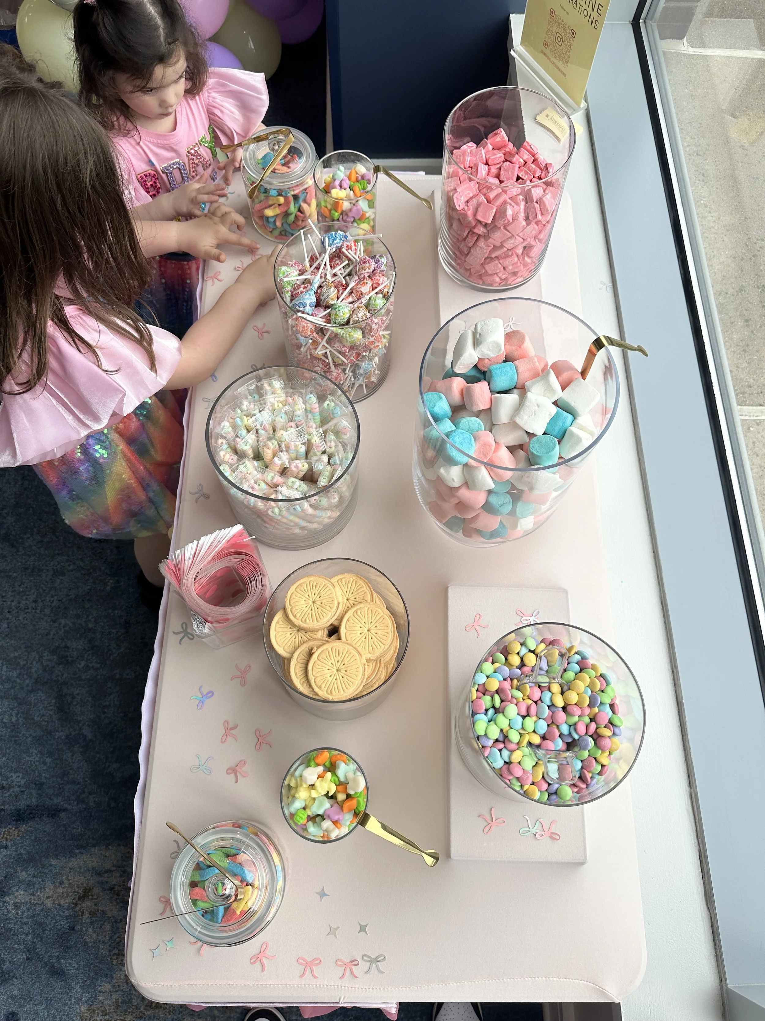 Children at a candy buffet with jars of colorful candies, marshmallows, marshmallow pops, and Cookies with pink napkins on a decorated table.