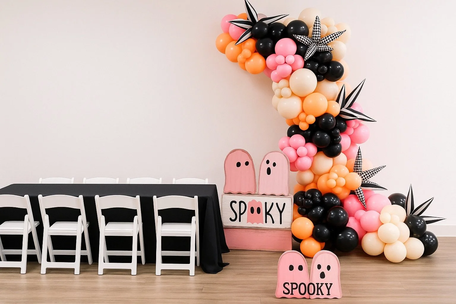 Decorative Halloween-themed balloon arch with pink, black, orange, and cream balloons, black and white striped leaves, and ghost cutouts with the word 'Spooky' on a pink box and on a pink ghost-shaped sign, next to a black table with white chairs.