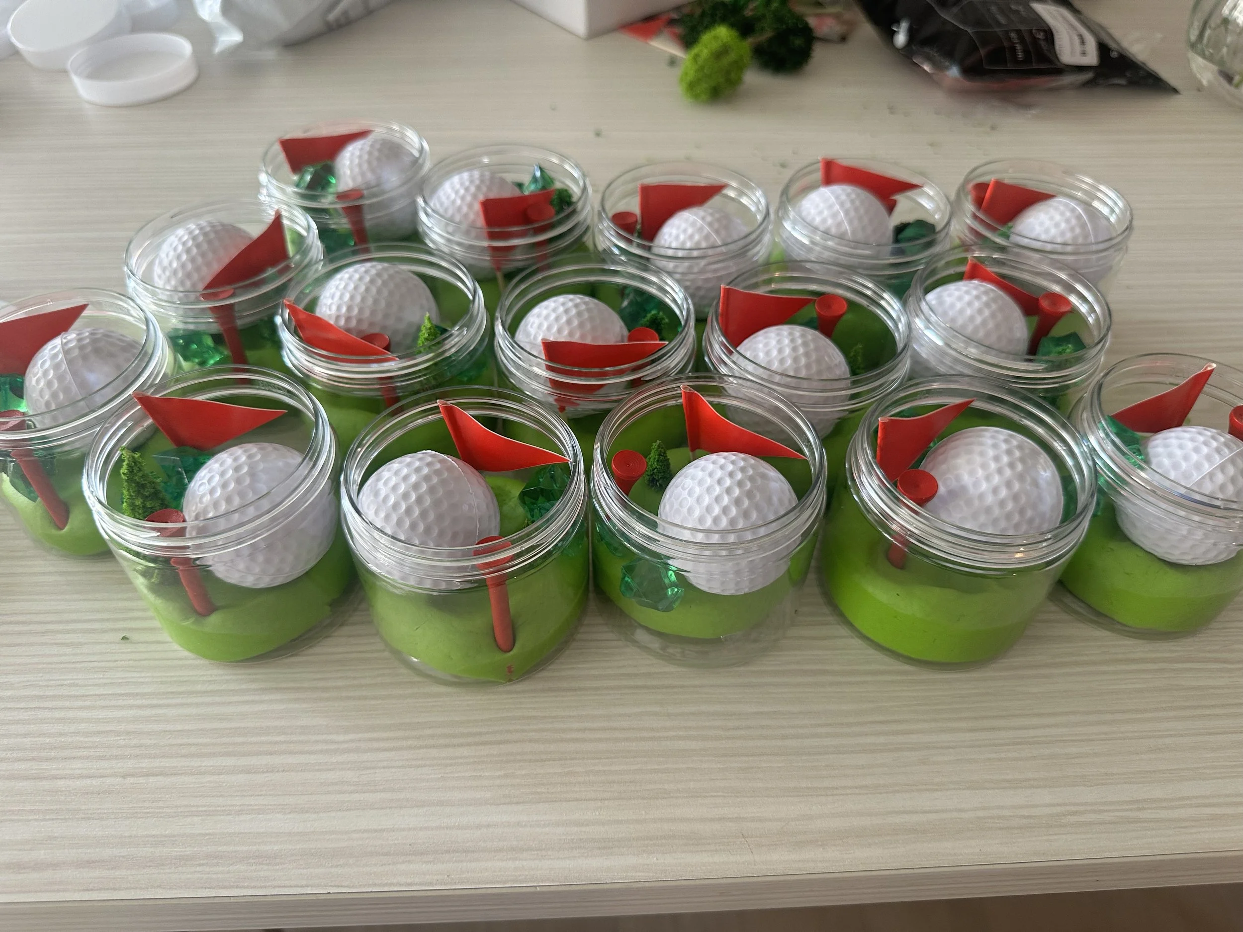 Multiple small containers filled with green clay, each topped with a white golf ball, red plastic flags, and small green and red decorative items, on a light-colored table.