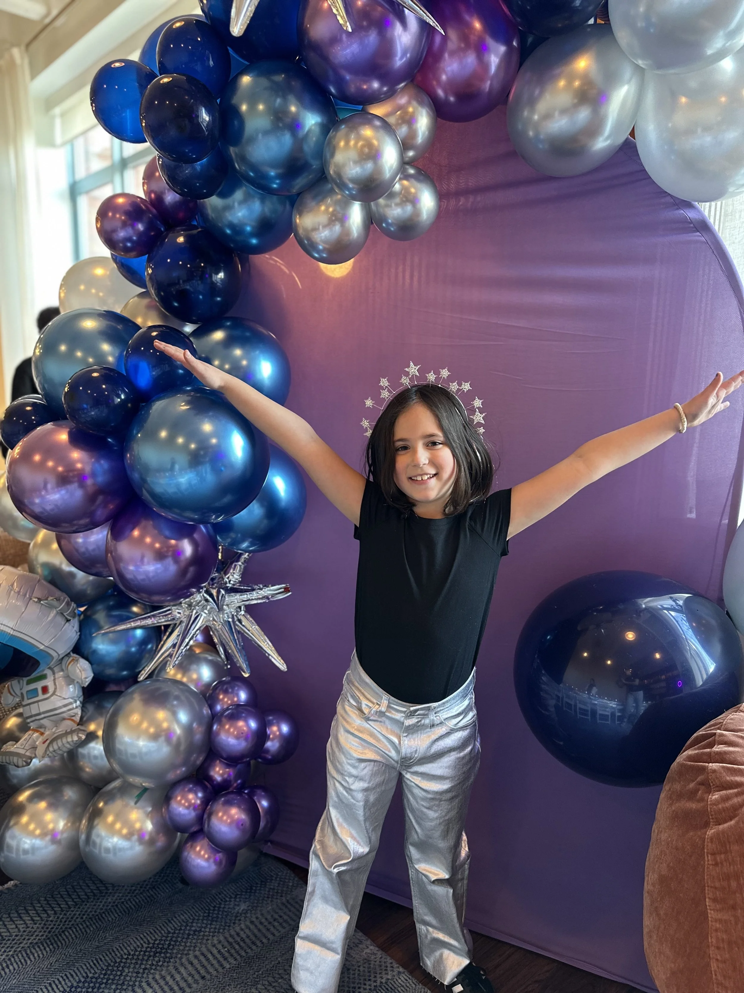 A young girl with shoulder-length dark hair wearing a black shirt and metallic silver pants, smiling with arms outstretched in front of a purple backdrop decorated with a large arrangement of purple, blue, and silver balloons. She also has a silver s