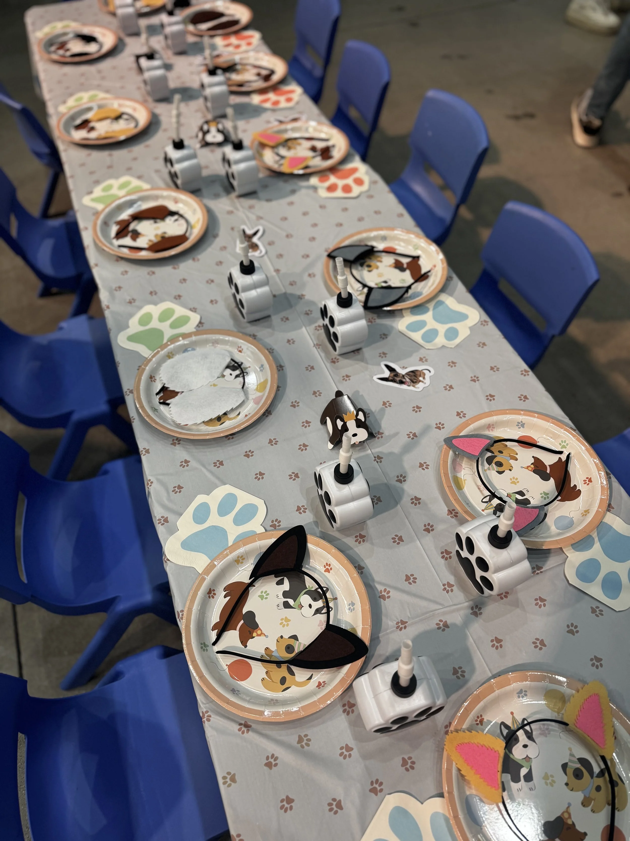 A long table decorated for a dog-themed party with plates, cup dispensers, and paper decorations featuring various cartoon dog faces.