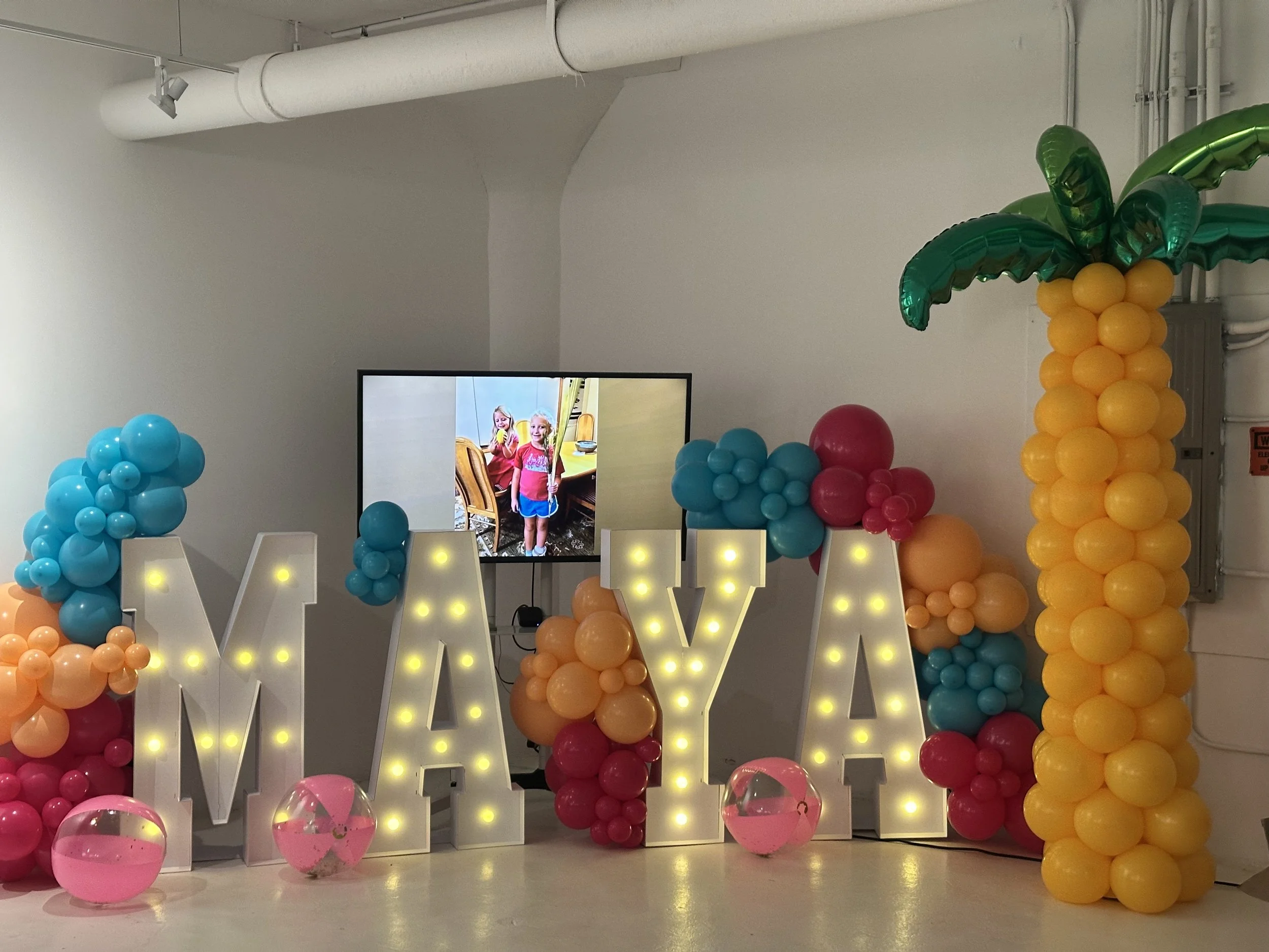 Decorations spelling out 'MAYA' with balloons of various colors including blue, pink, orange, red, and yellow, along with illuminated marquee letters, a tropical banana-shaped balloon column, and a monitor displaying a photo of two young girls.