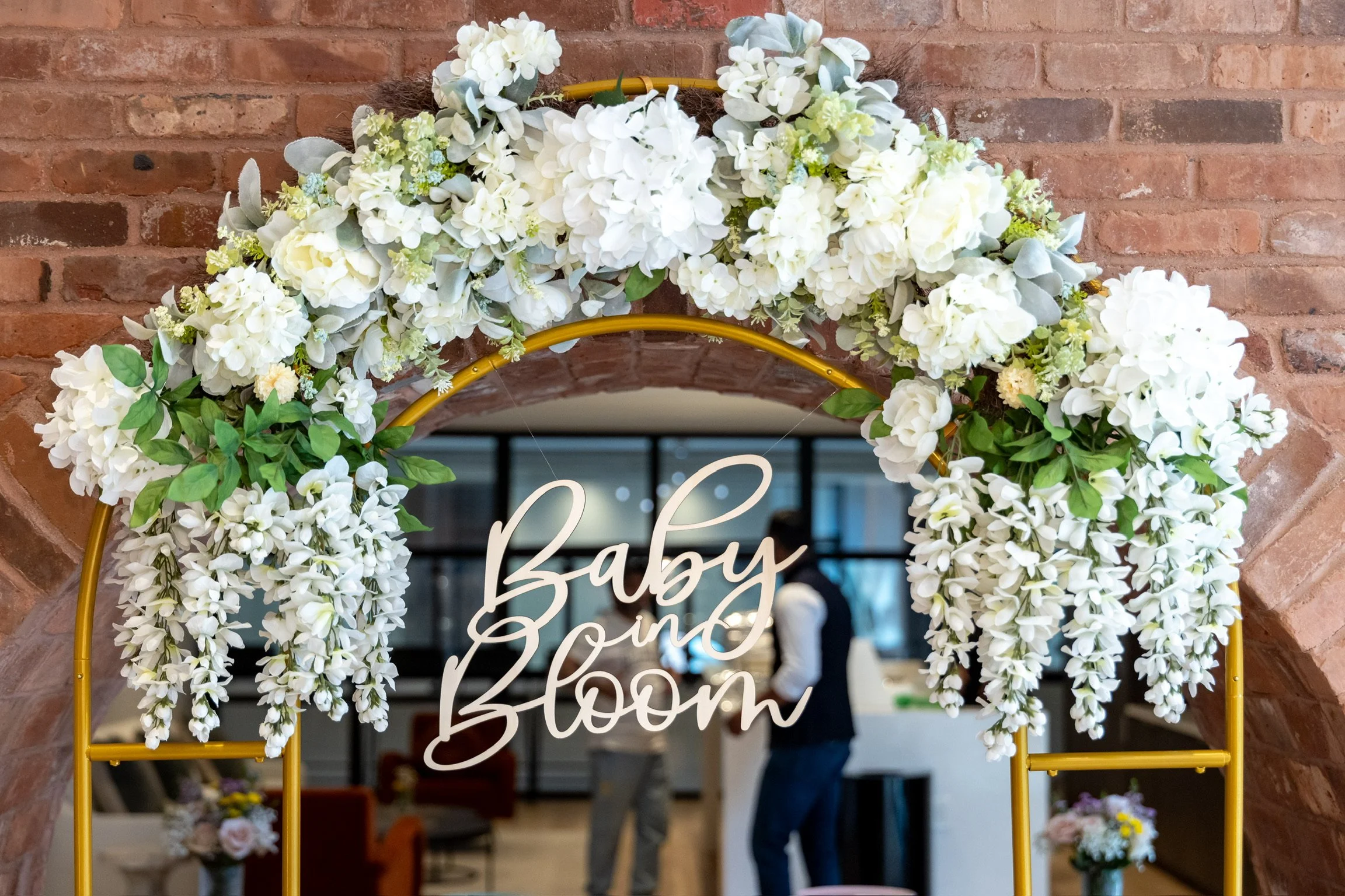 Floral arch decorated with white flowers and green leaves, with a sign that reads "Baby Bloom" hanging in the center. Indoor setting with a brick wall background and blurred people in the background.