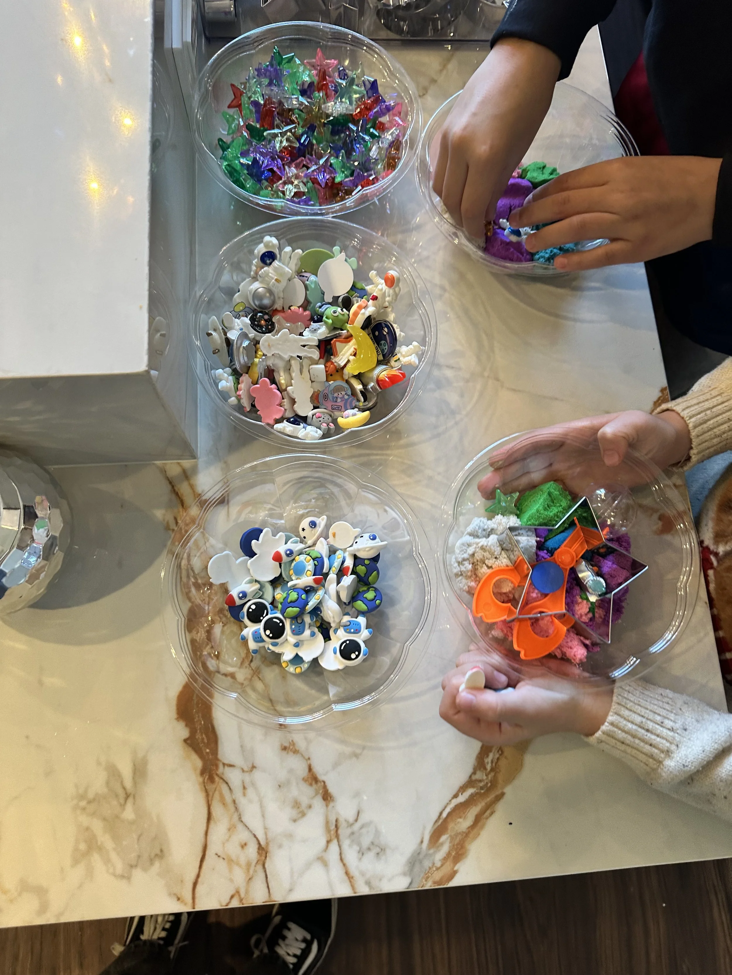 People's hands reaching into clear containers filled with colorful foam and small figurines on a marble table.