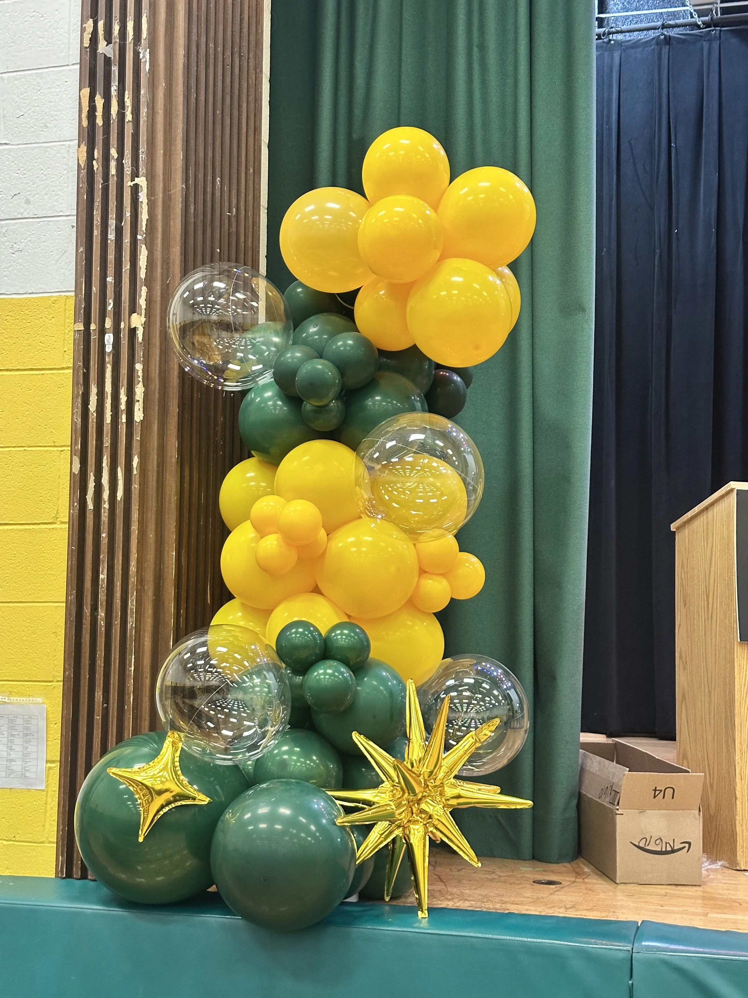 Decorative balloon arrangement with green, yellow, and transparent balloons and gold star-shaped balloons, set against a stage with curtains.