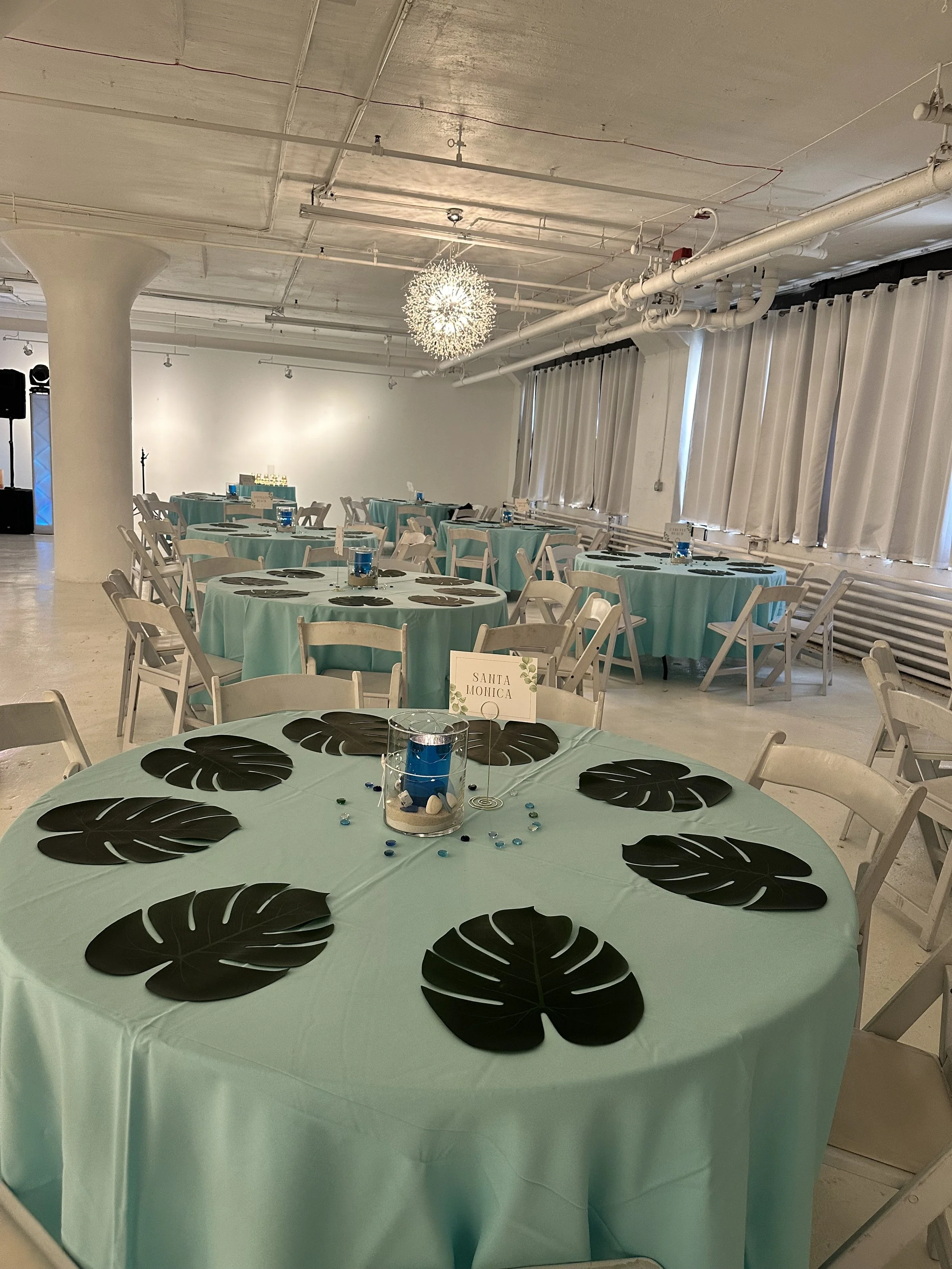 Up close view of a round table with a light blue tablecloth, decorated with six black monstera leaf placemats, a centerpiece with a blue candle, and a small sign labeled 'Santa Monica.' The table is set with black napkins and surrounded by white chai
