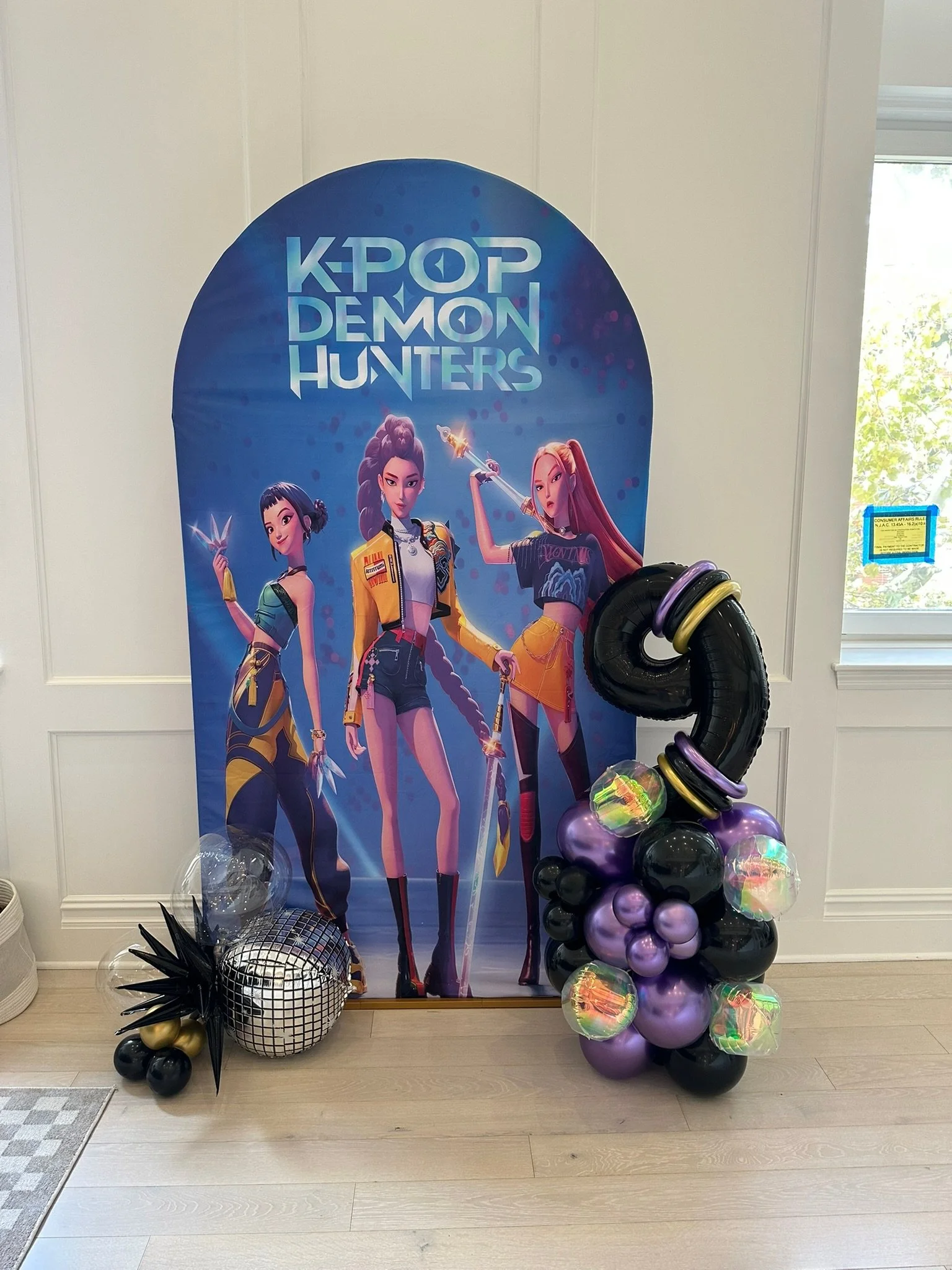 K-pop Demon Hunters poster with three animated female characters in colorful outfits, next to balloon arrangements including a silver disco ball, black spiked balloon, and purple and black balloons with iridescent accents.