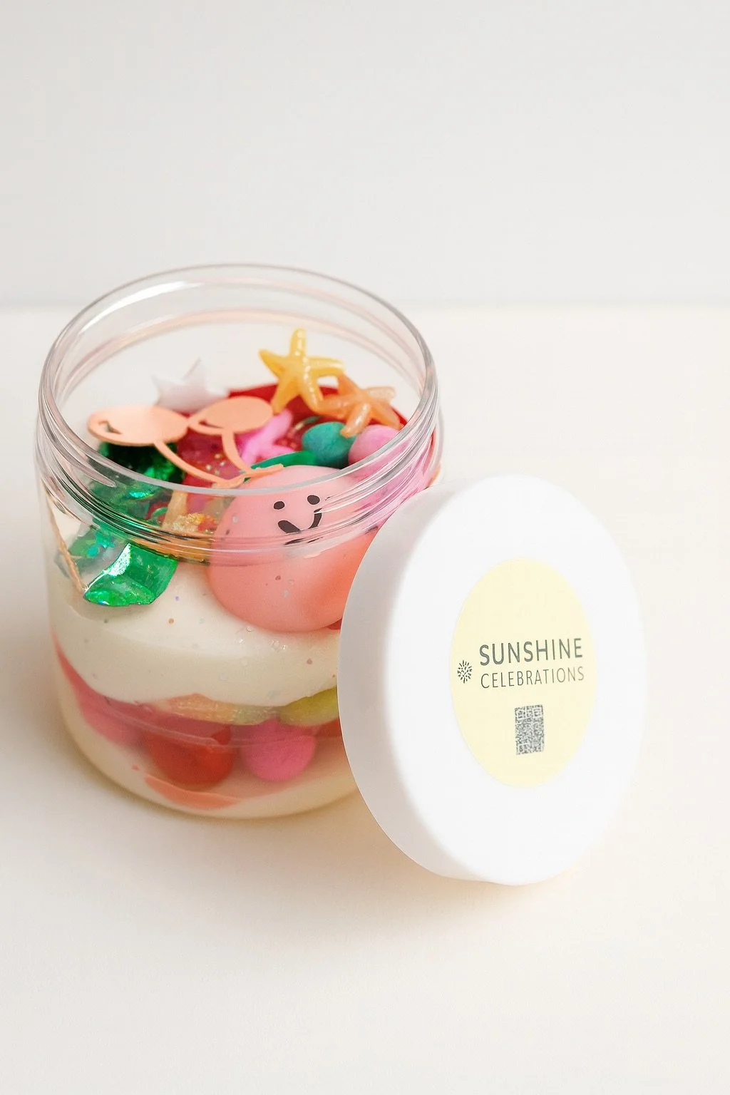 Colorful layered dessert in a glass jar with decorative sweets inside, and a white lid labeled 'Sunshine Celebrations' placed beside it.