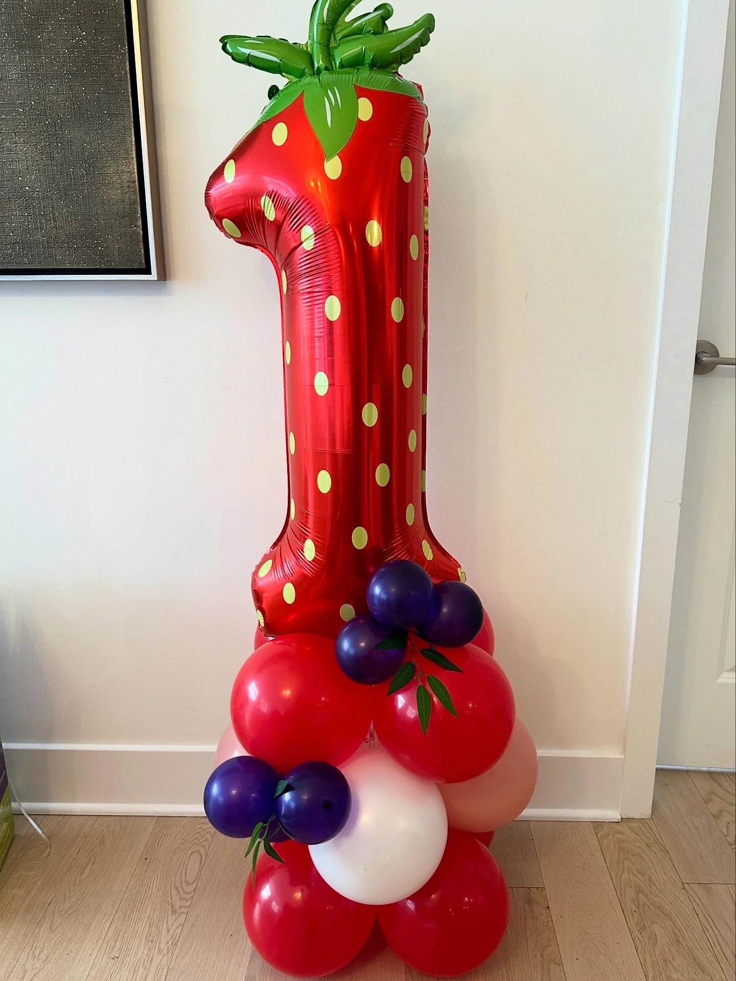 A large balloon sculpture resembling a strawberry plant, with a tall, red, polka-dotted balloon as the main stem, topped with green balloon leaves, and decorated with smaller purple, red, pink, and white balloons at the base.