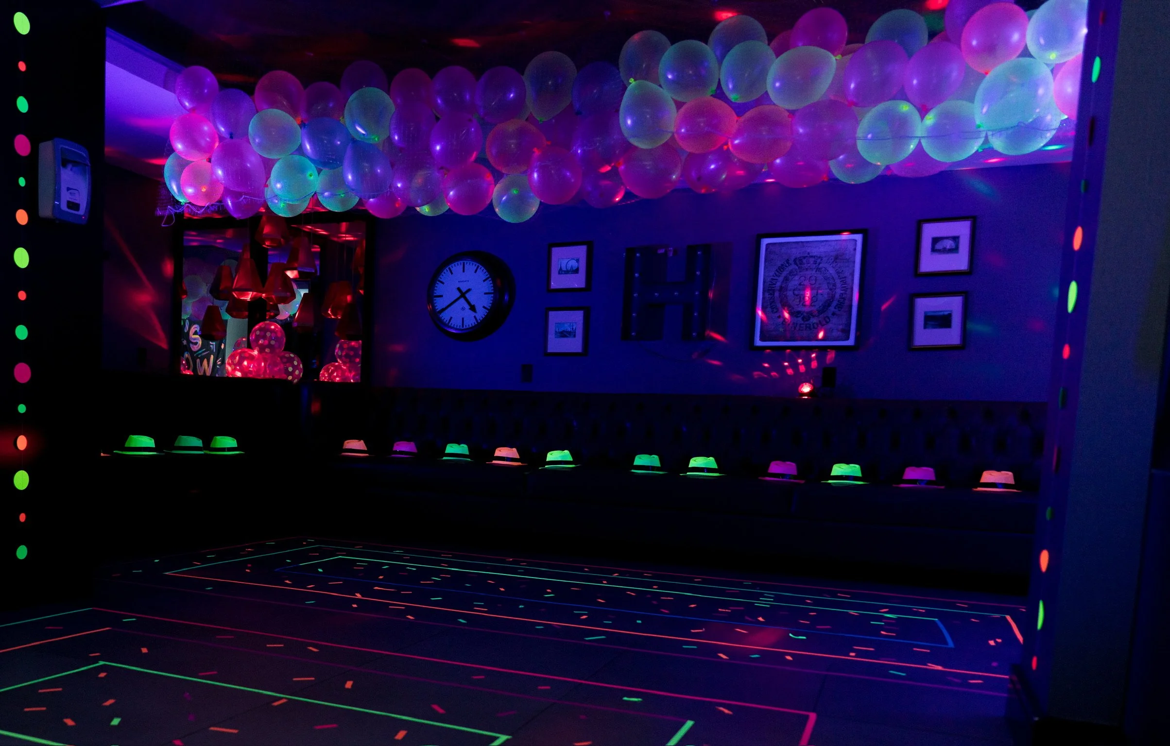 A dimly lit indoor space decorated with blacklights, neon-colored balloons on the ceiling, glowing hats on a bench, a wall clock, framed pictures, and laser light patterns on the floor and walls, creating a party or celebration atmosphere.