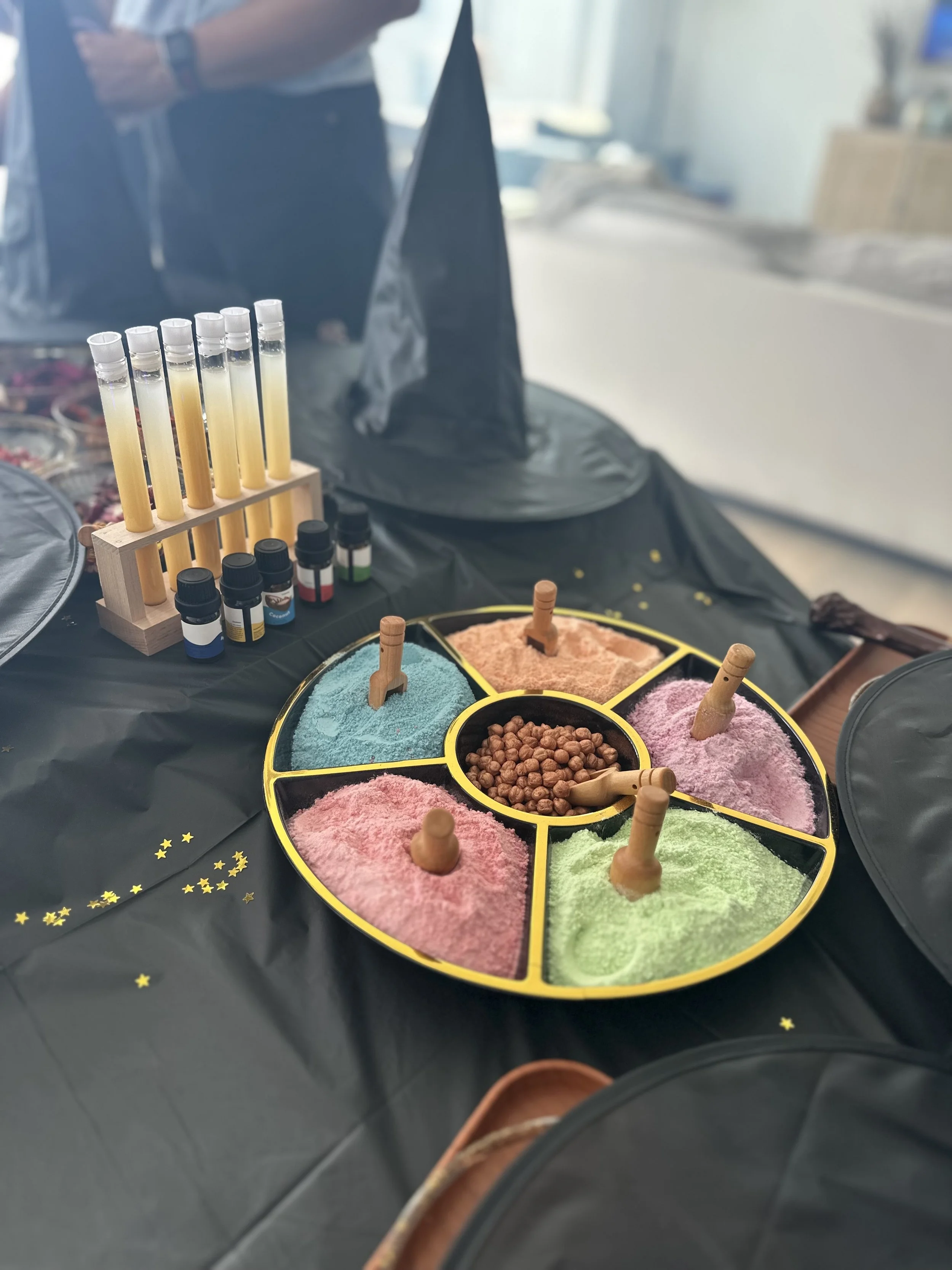 Colorful salt or sand in small bowls with wooden scoops, arranged around a circular tray, with test tubes containing yellowish liquids and small bottles of essential oils or flavorings in the background. The setup appears to be for a sensory, baking,