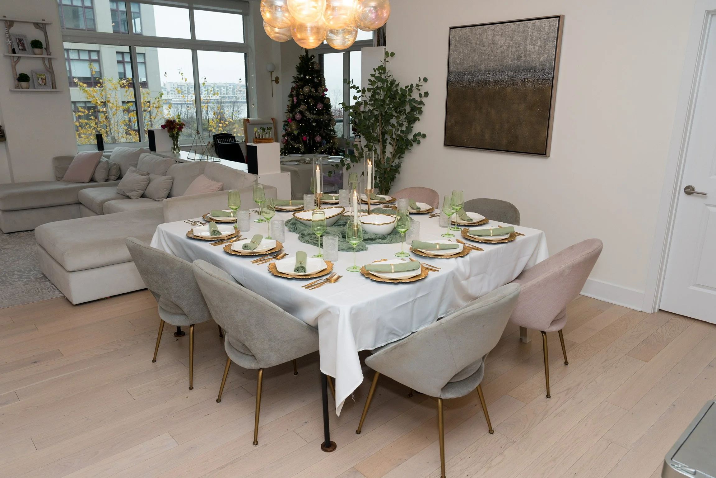 Dining table set with green glassware, gold charger plates, and cloth napkins in a modern living space decorated for Christmas with a Christmas tree, a large abstract painting on the wall, and a chandelier with glass globes.