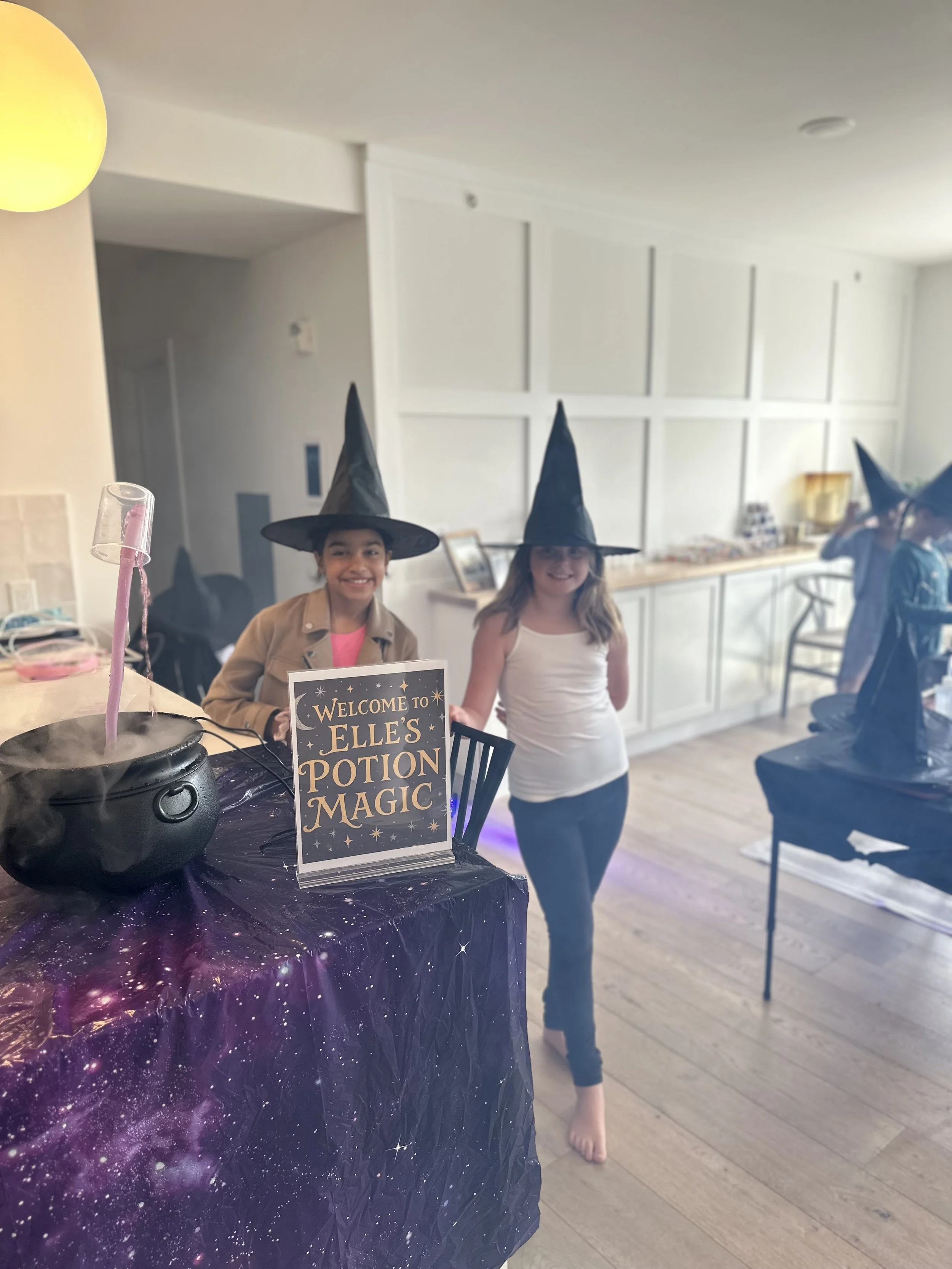 Two girls wearing witch hats at a potion magic themed party, standing behind a table with a sign that says "Welcome to Elle's Potion Magic," with a black cauldron emitting smoke and a purple galaxy themed tablecloth.
