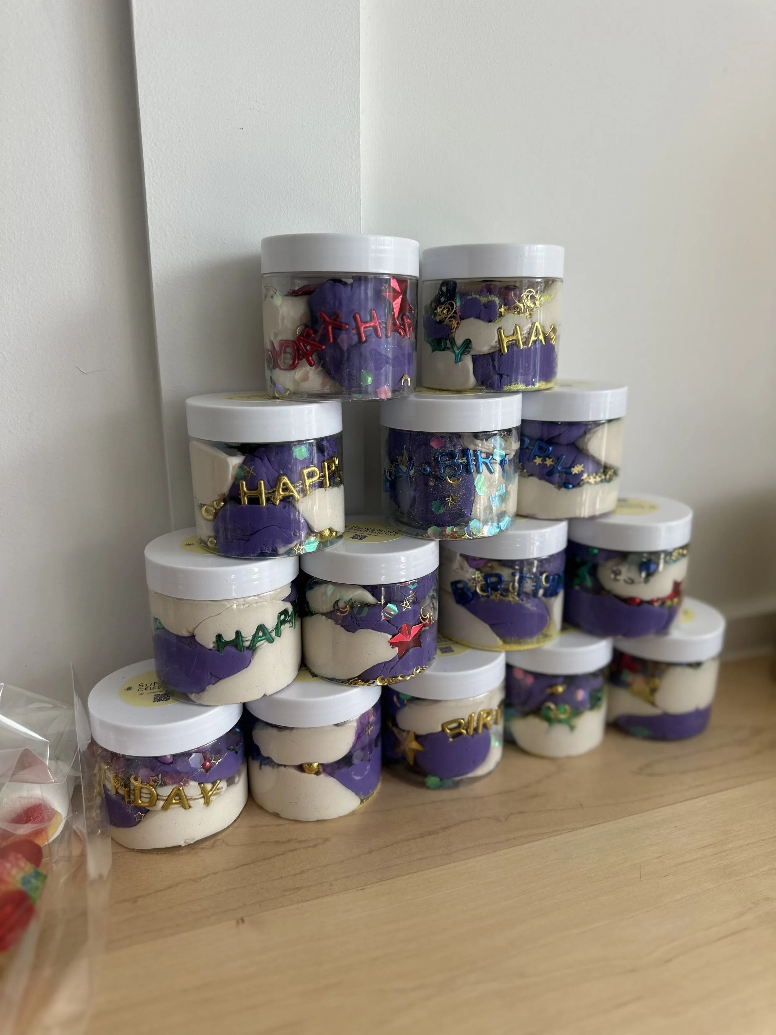 Stacked plastic jars filled with white and purple slime, decorated with colorful glitter and 'Happy Birthday' and 'Birthday' letter charms.