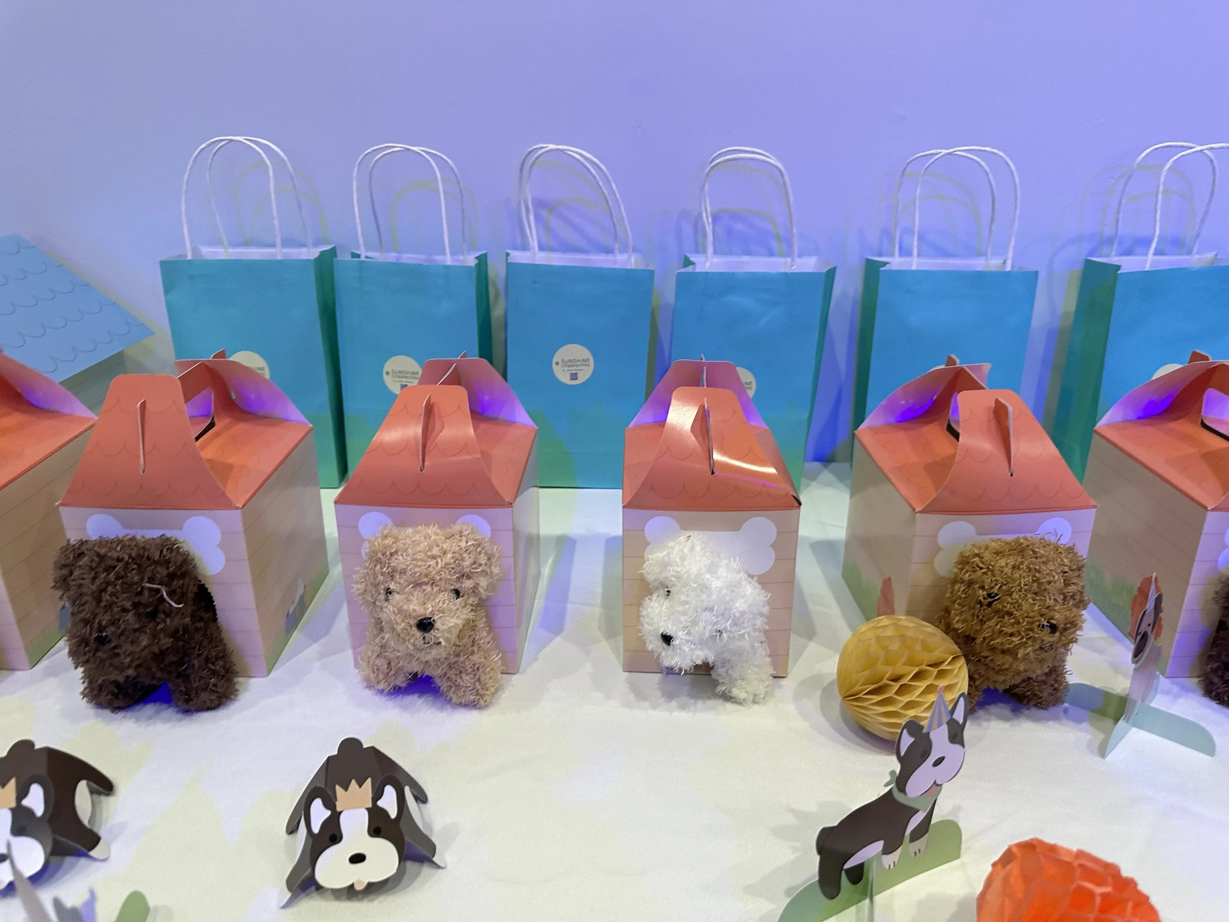 Display of small plush puppy toys, dog-themed party favor boxes with pink roofs, blue gift bags with white handles, decorative dog cutouts, and honeycomb paper balls on a table.