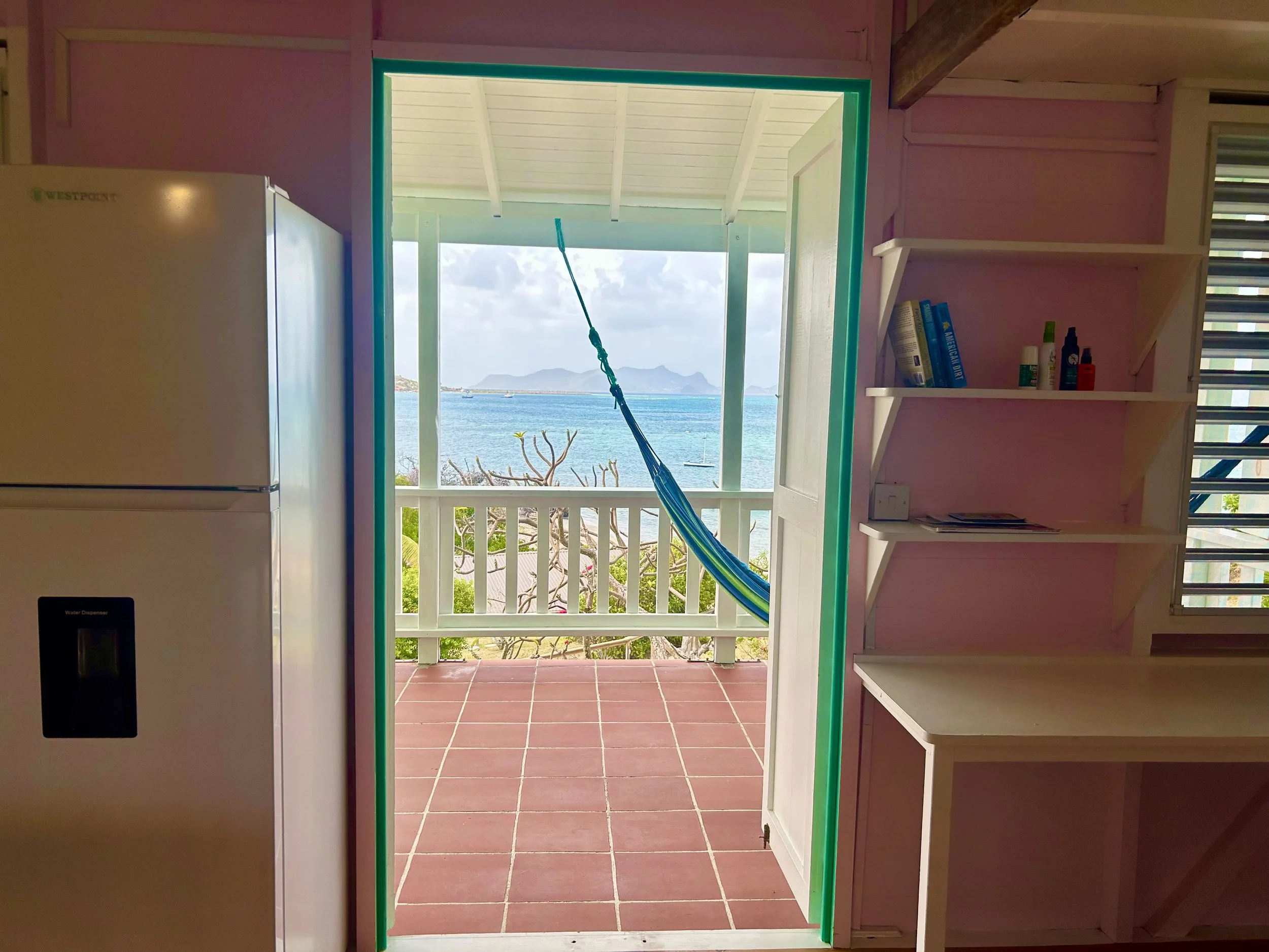 View through an open door revealing a balcony with a hammock overlooking the ocean and islands in the distance, interior includes a refrigerator and shelves with books and bottles.