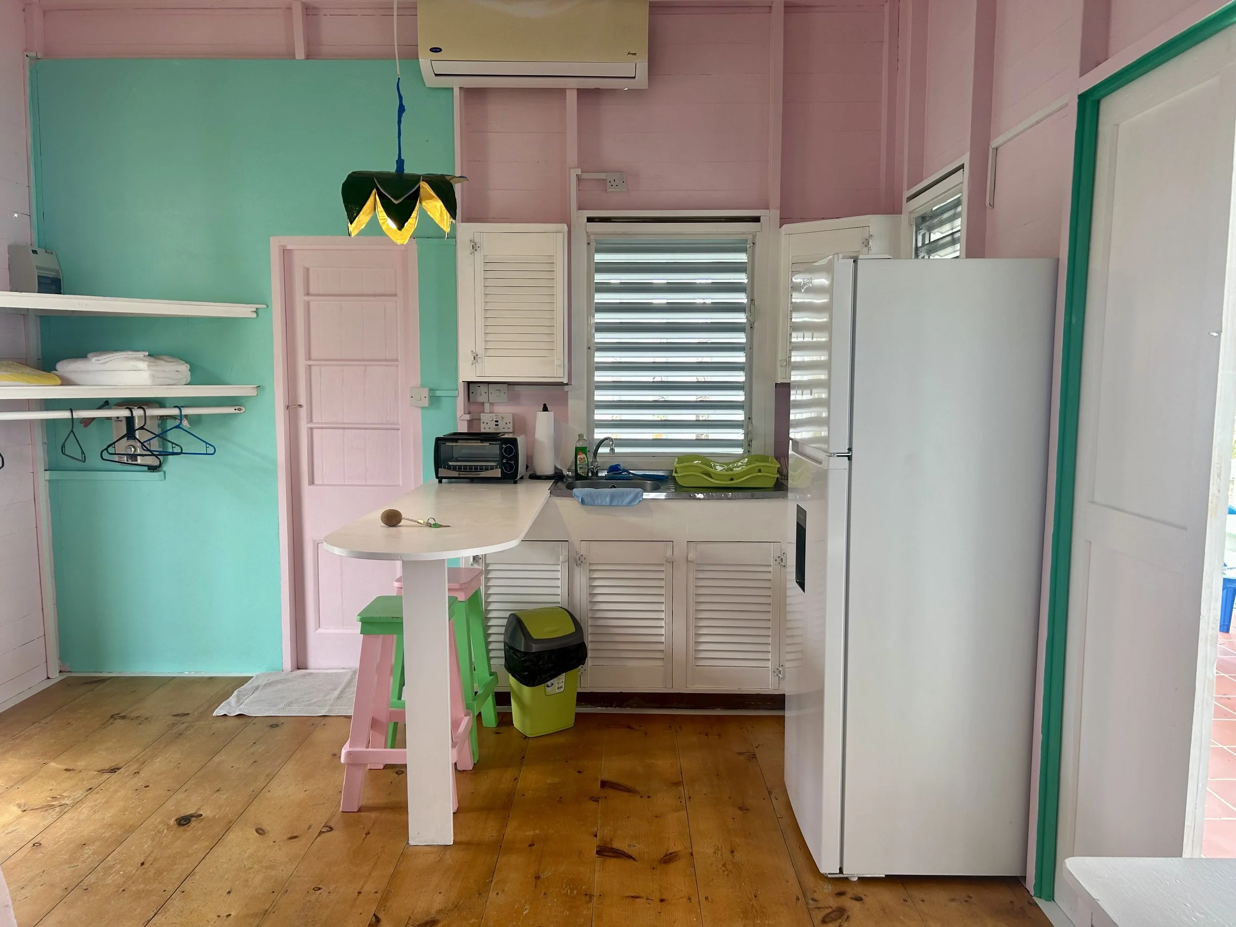 A small kitchen with pastel pink and turquoise walls, white cabinets, a refrigerator, a window with louvers, and a wooden floor. There is a countertop with a toaster and a sink, and a small breakfast bar with a pink and green stool.