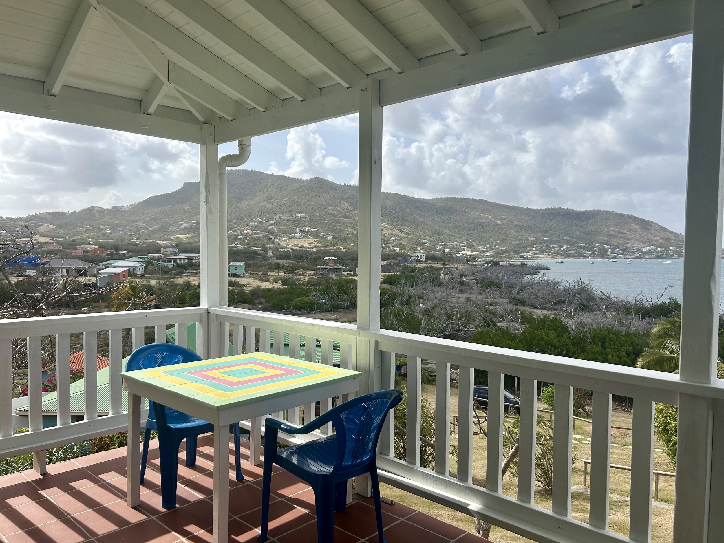 View from a balcony overlooking a coastal landscape with mountains, houses, trees, and water, featuring a colorful table and two blue chairs.