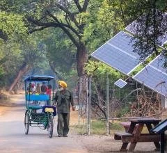 A person riding a rickshaw while another walks along a dirt path in a rural setting with trees and solar panel structures in the background.