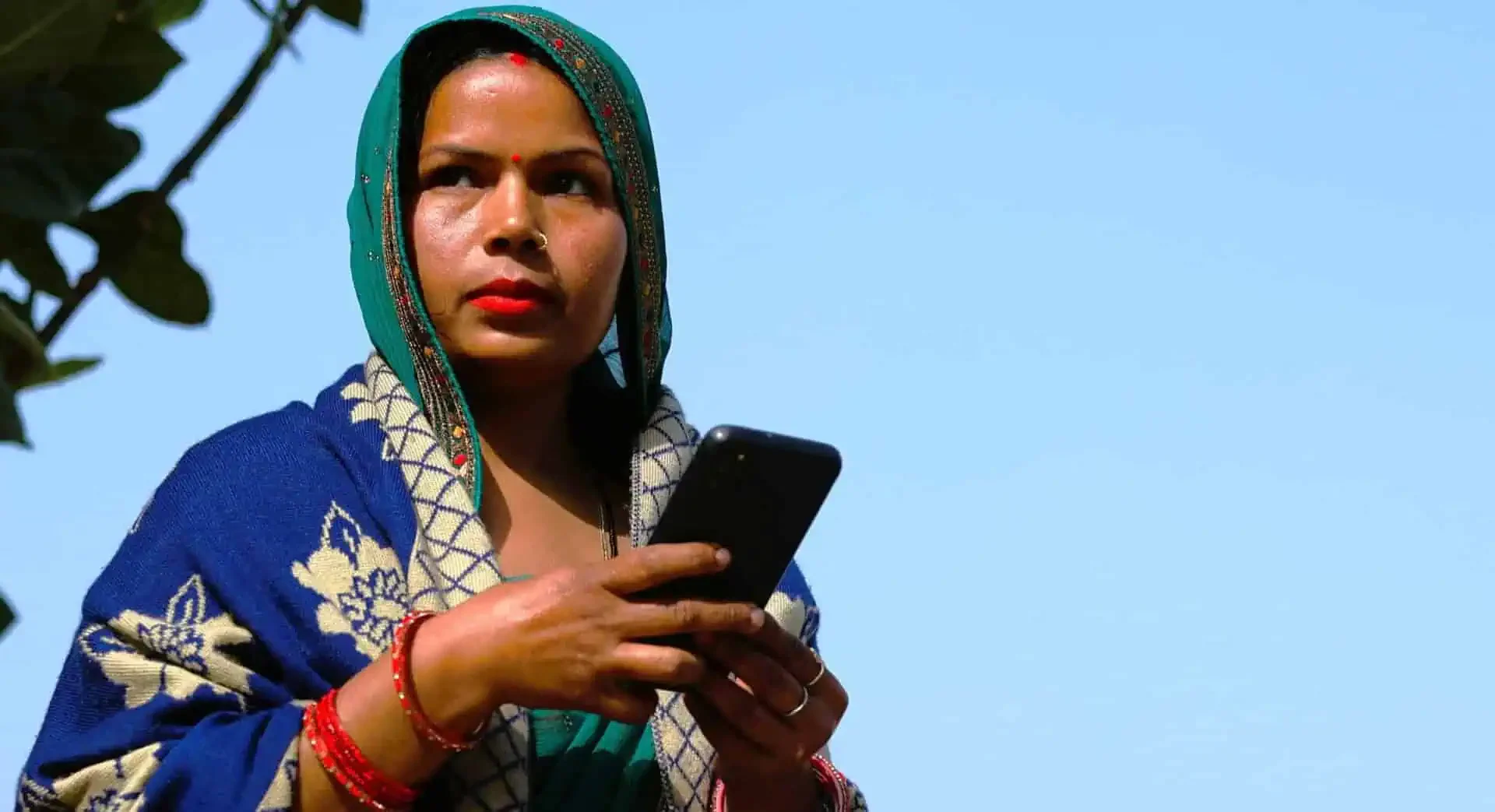 A woman wearing traditional blue and green attire with intricate embroidery, red bangles, and a red bindi on her forehead, holding a smartphone and standing outdoors against a clear blue sky.
