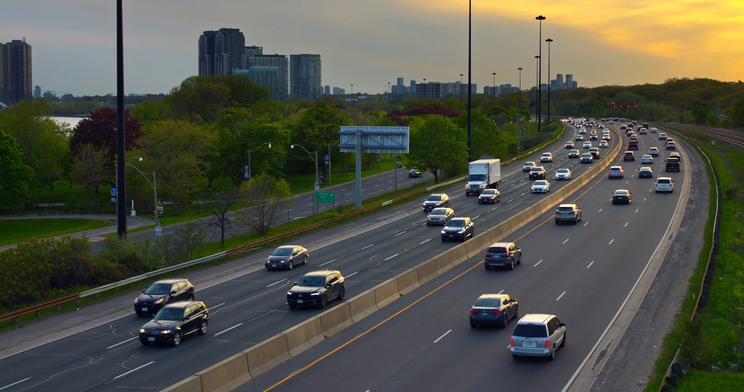 Autoroute avec beaucoup de voitures, entourée d’arbres verts, avec un skyline de bâtiments modernes en arrière-plan, au coucher du soleil.