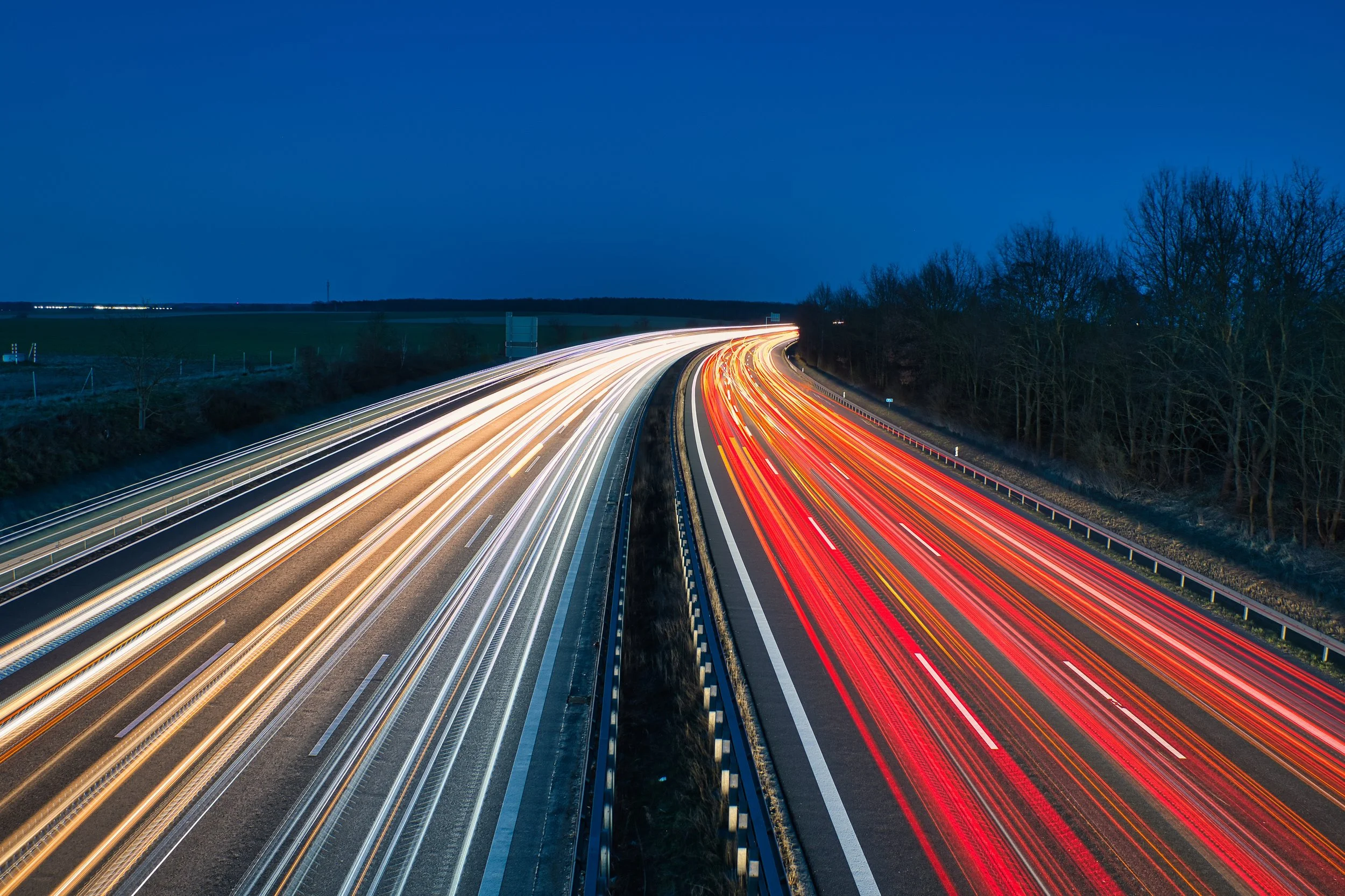 Longue exposition photographiant une autoroute la nuit, montrant des traînées lumineuses de véhicules rouge et blanc, avec un ciel sombre et des arbres à proximité.